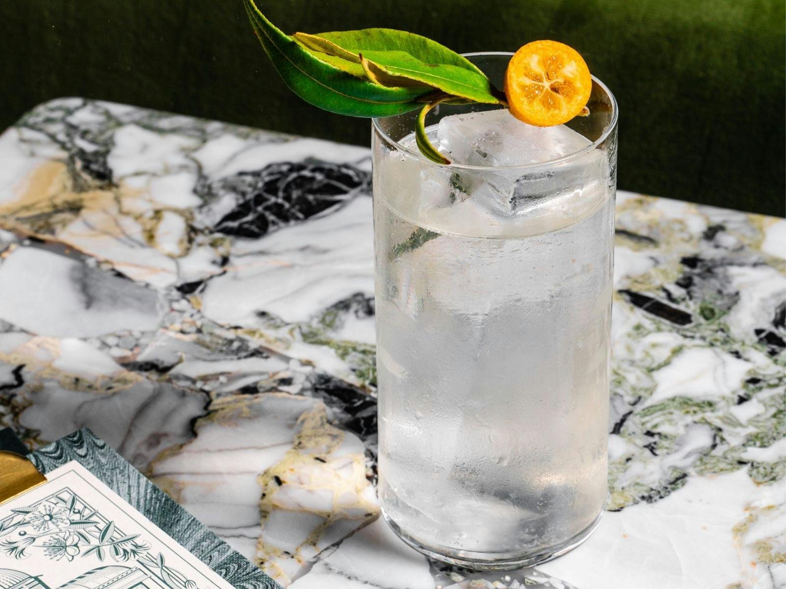 Clear cocktail in tall glass garnished with kumquat