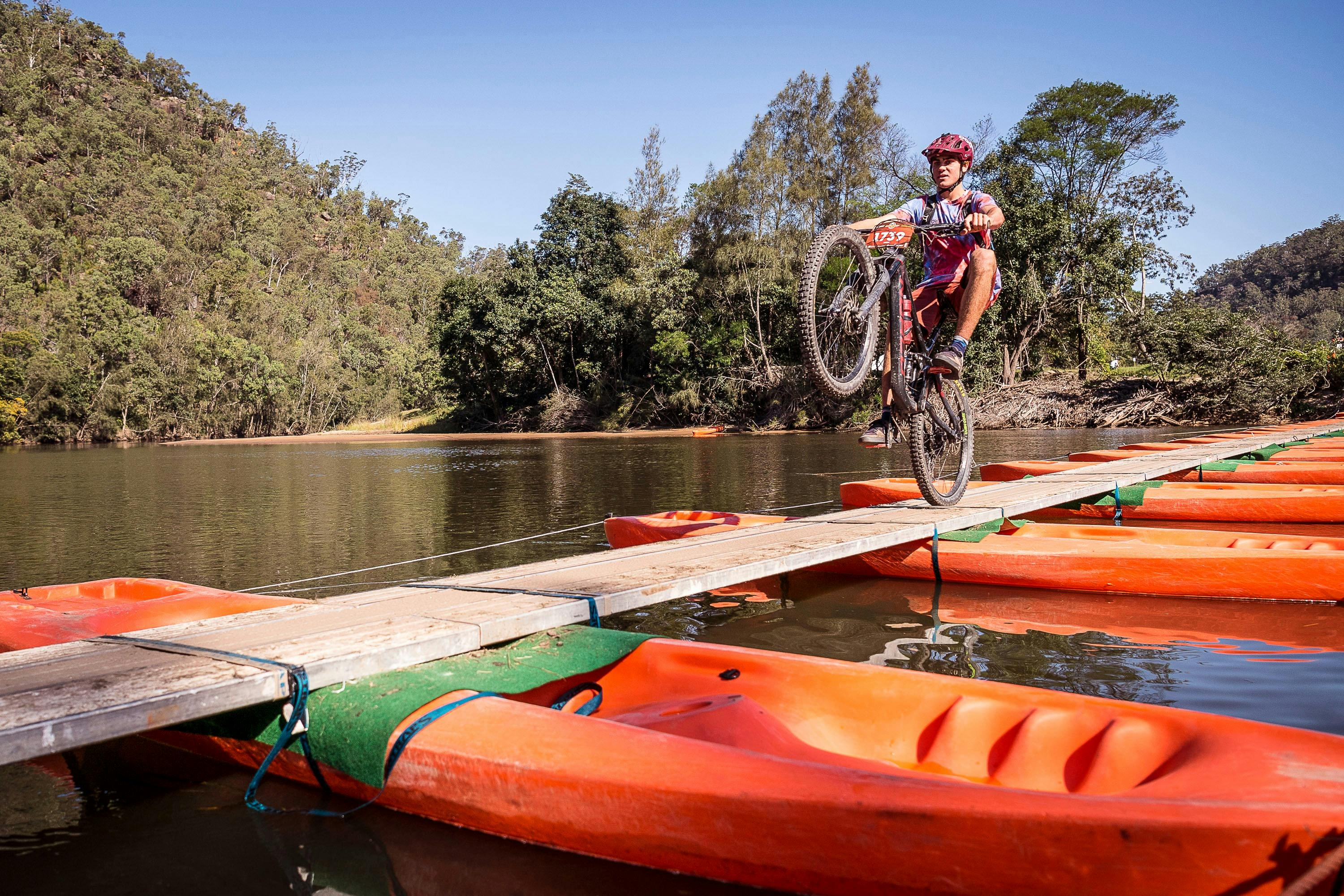 Mountain bike rider performs wheelie while on kayak bridge across the river.