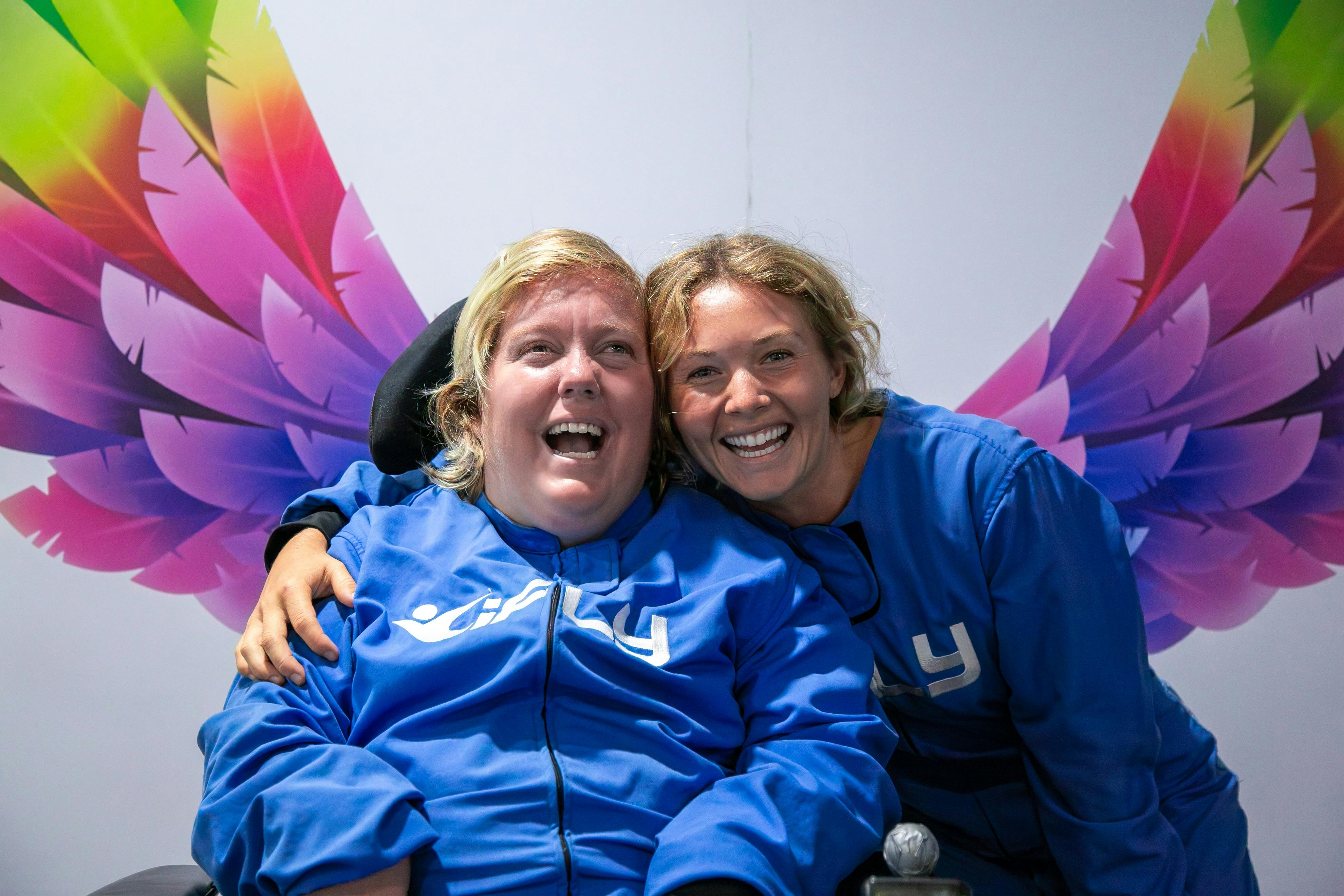 Happy flyer who uses a wheelchair with her carer in front of a wings graphic.
