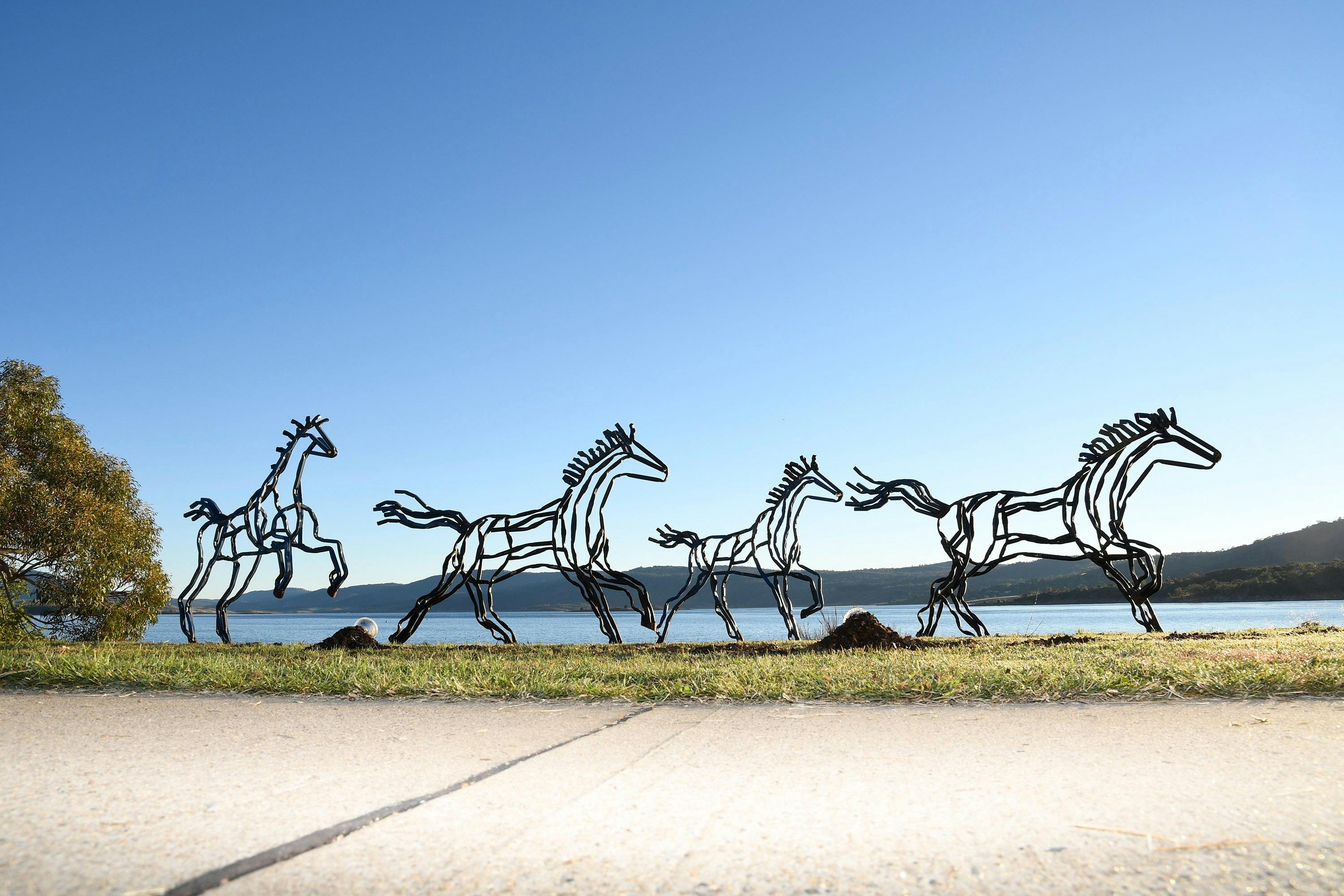 Metal horse sculptures in motion by a lake, set against a clear blue sky.