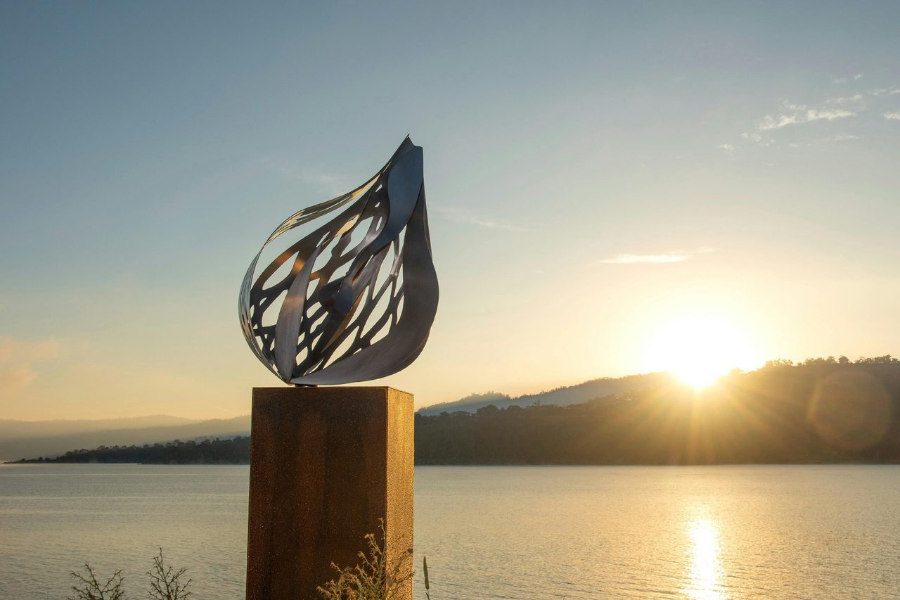 Metal sculpture of an intricate pod on a plinth, set against a still lake at sunrise.