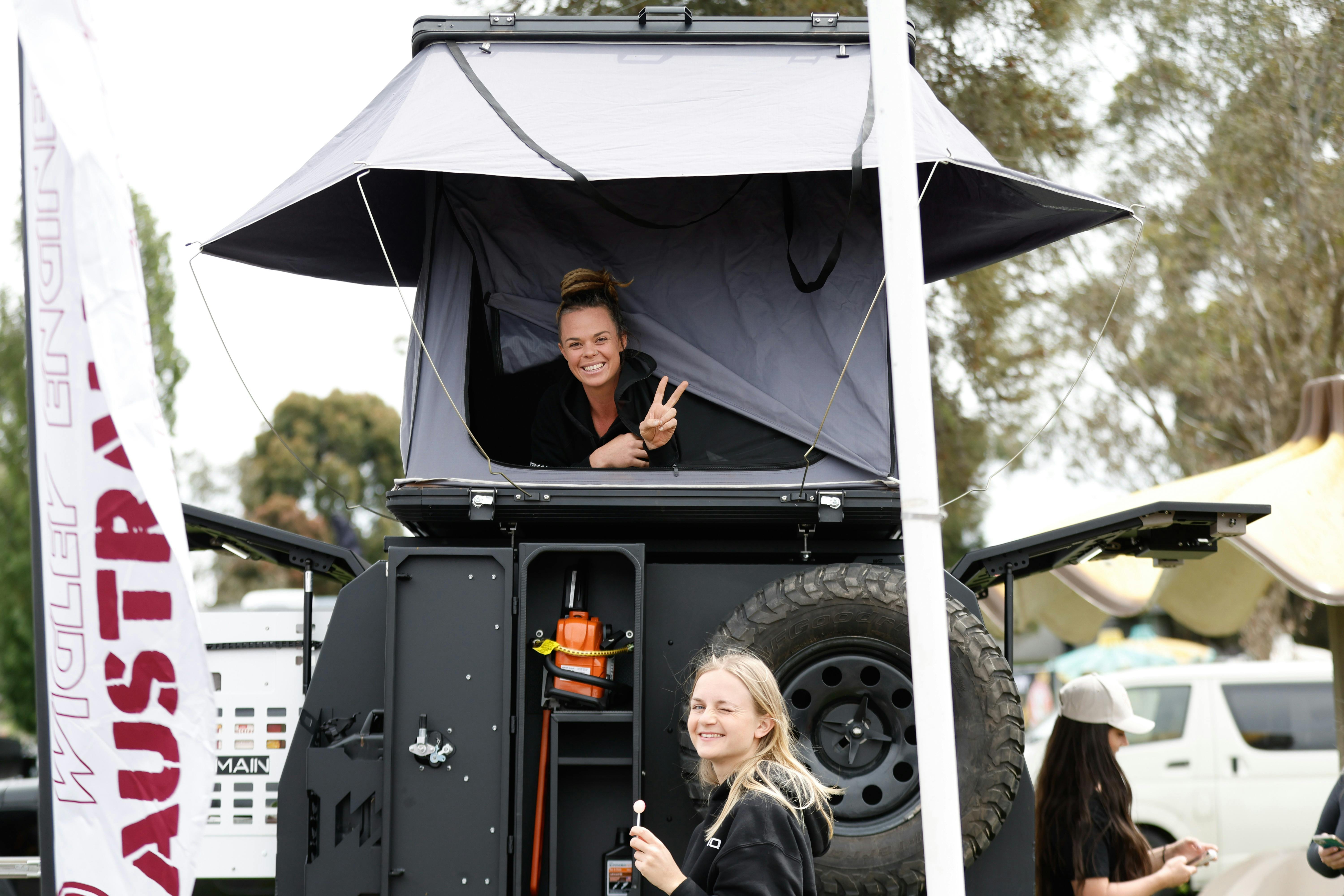 One young women lying in rooftop tent giving peace sign and  a second down below with a lollipop