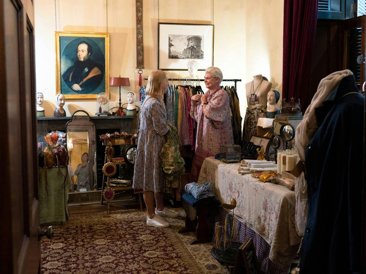 Two women chat infront of a clothing rack. Historic art hangs on the walls.