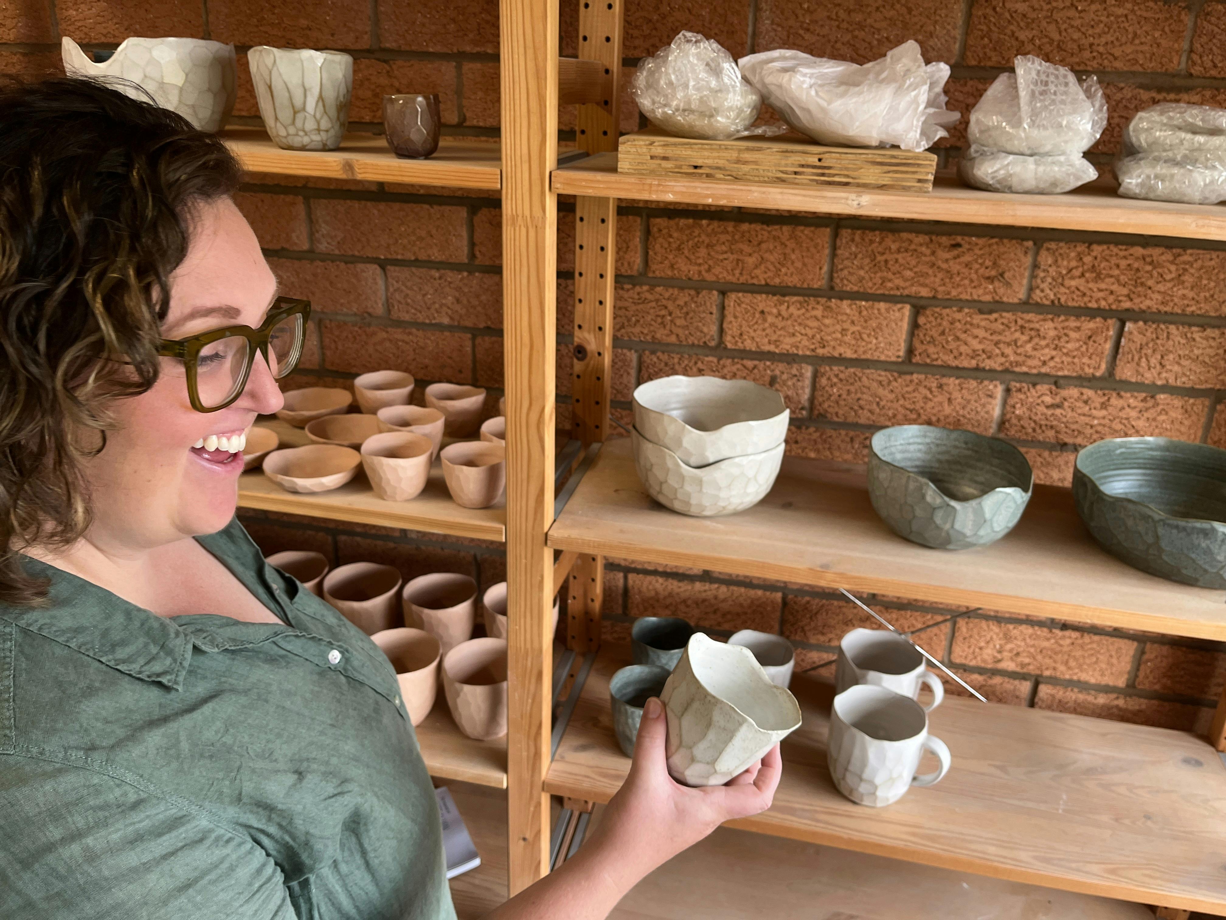 Woman with green shirt and glasses smiles while looking at cup in her hand shelves of pots behind