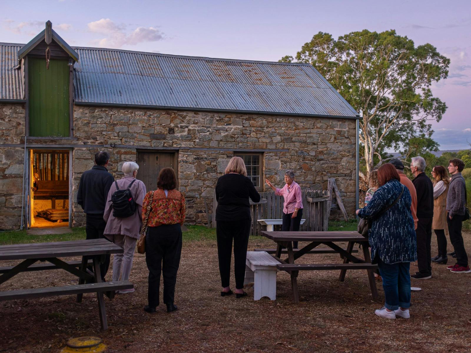 People gather out the front of a stone stable. The light spills out from the doorway as the sun sets