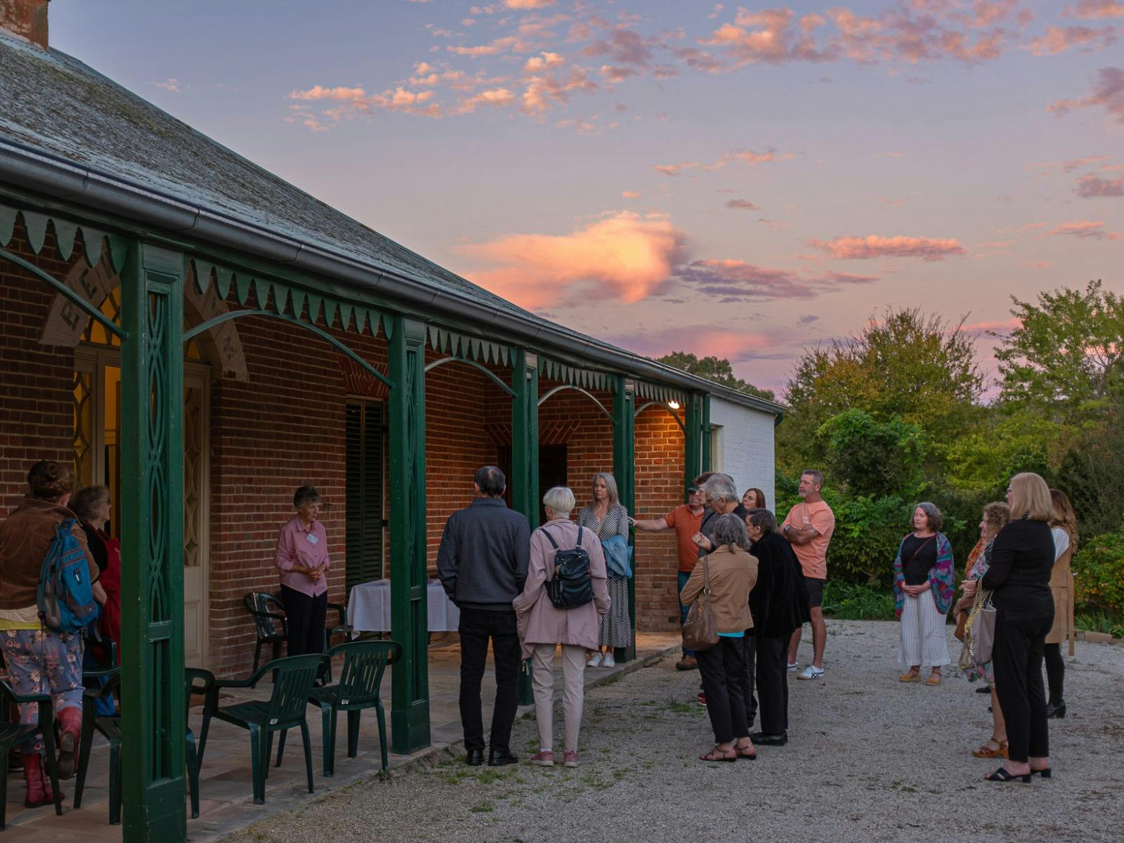 People gather on the verandah of a historic red brick house. The sun sets above.