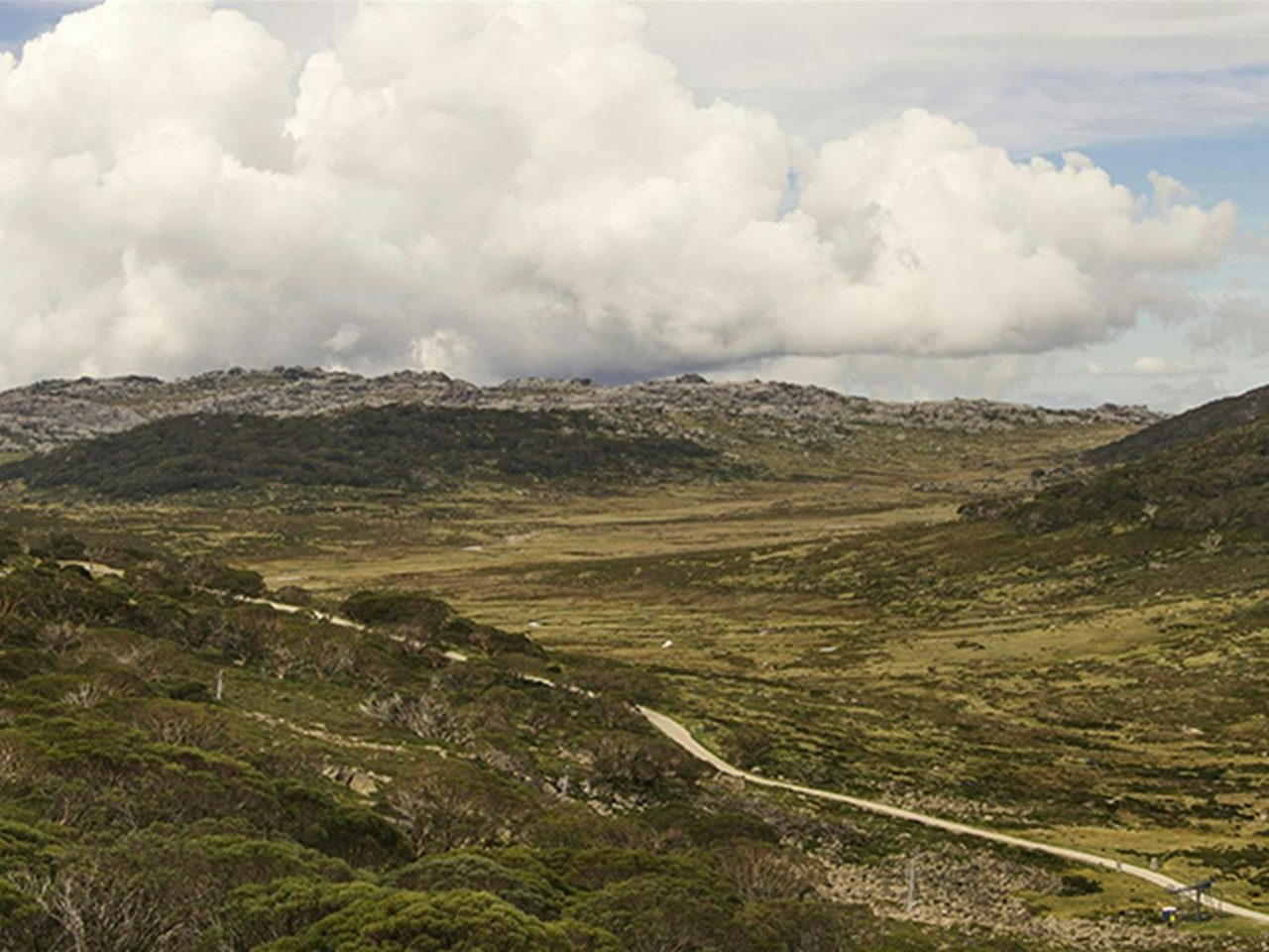 Wide view of open, rugged mountain landscape traversed by a dirt track, under partly cloudy skies.