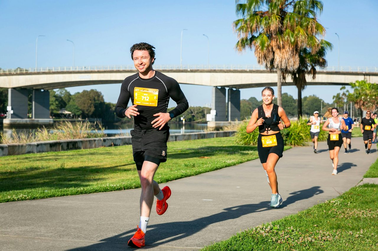 Two runners smiling running  past a bridge along the Parramatta River