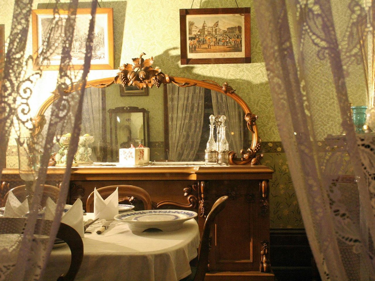 View peers through lace curtains to see a mirror upon drawers and a dining table, lit by lamplight