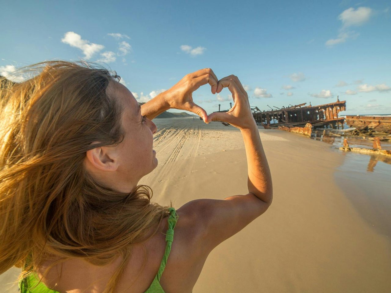 A young blonde woman holds her fingers in a love heart sign at a shipwreck on a sandy beach