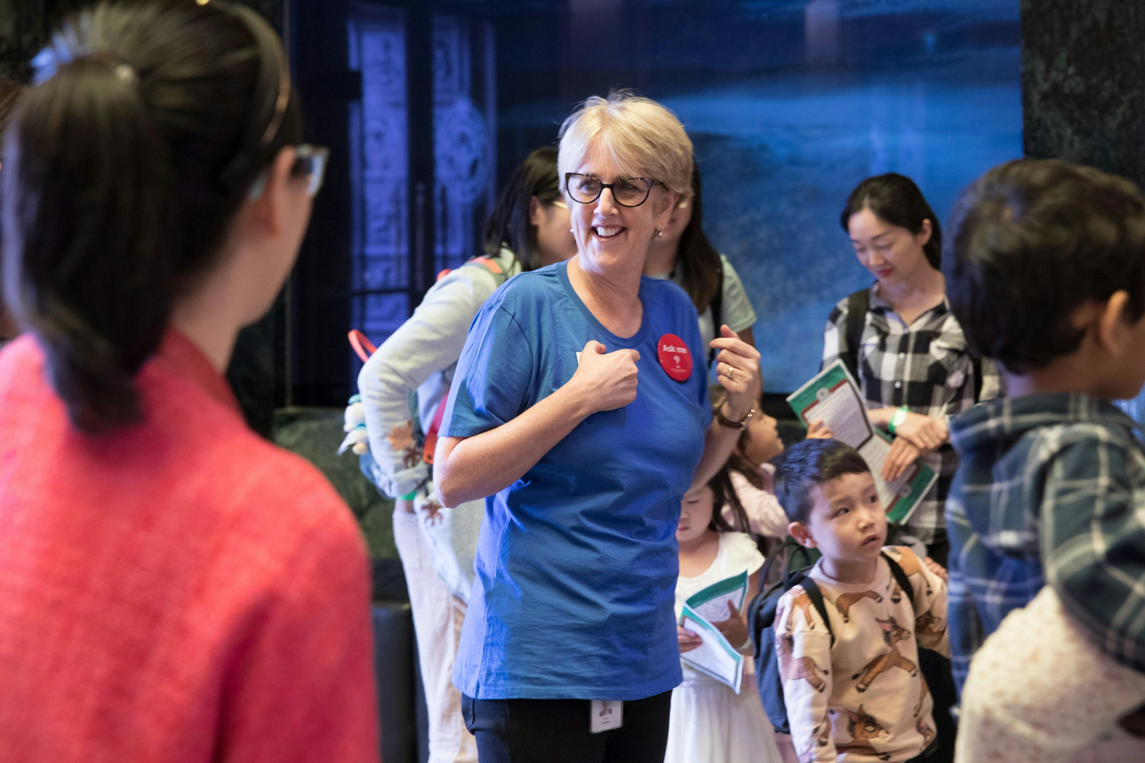 Woman wearing an 'Ask me' badge in the foyer of a Library helping a group of children and parents