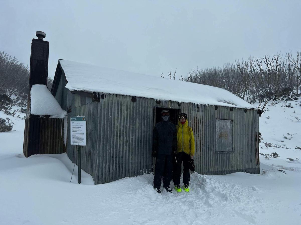 Two hikers standing outside a hut in the snow.