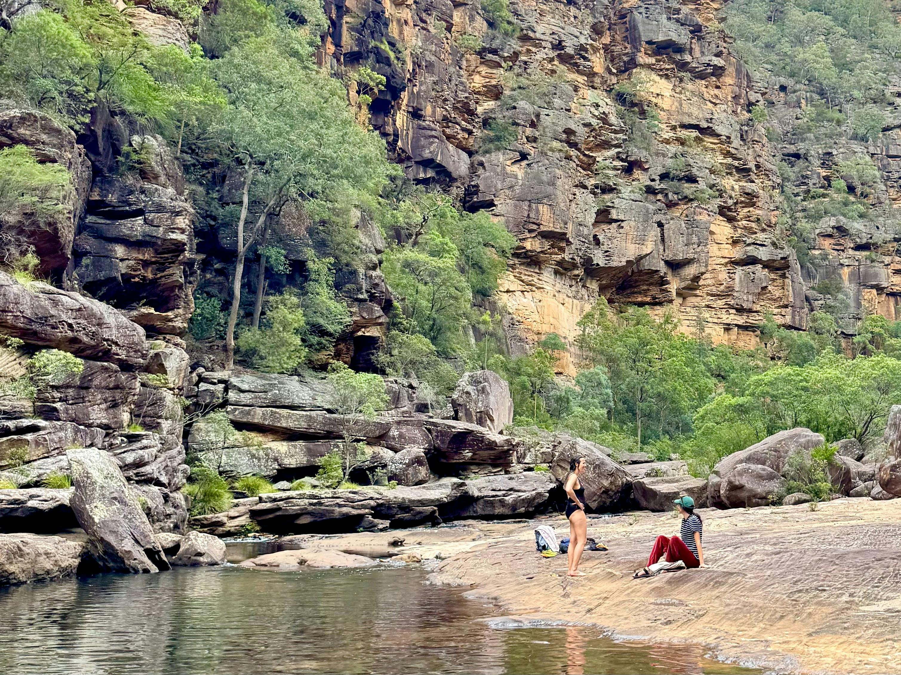 2 women chat next to the water, with sheer cliffs behind them