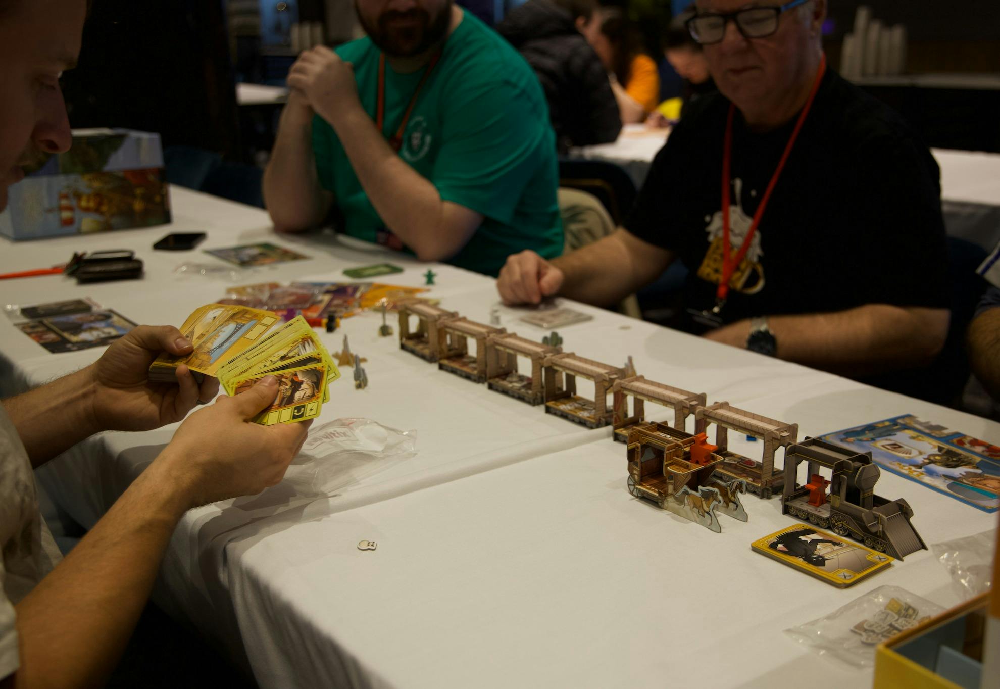 Three men sit around a table playing a board game with cards in their hand and a cardboard 3D train