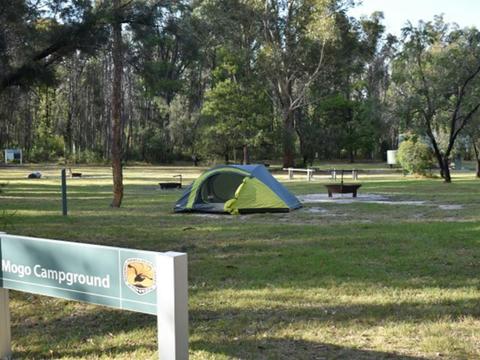 A sign at Mogo campground with a tent and woodfire barbecues in the background at Yengo National