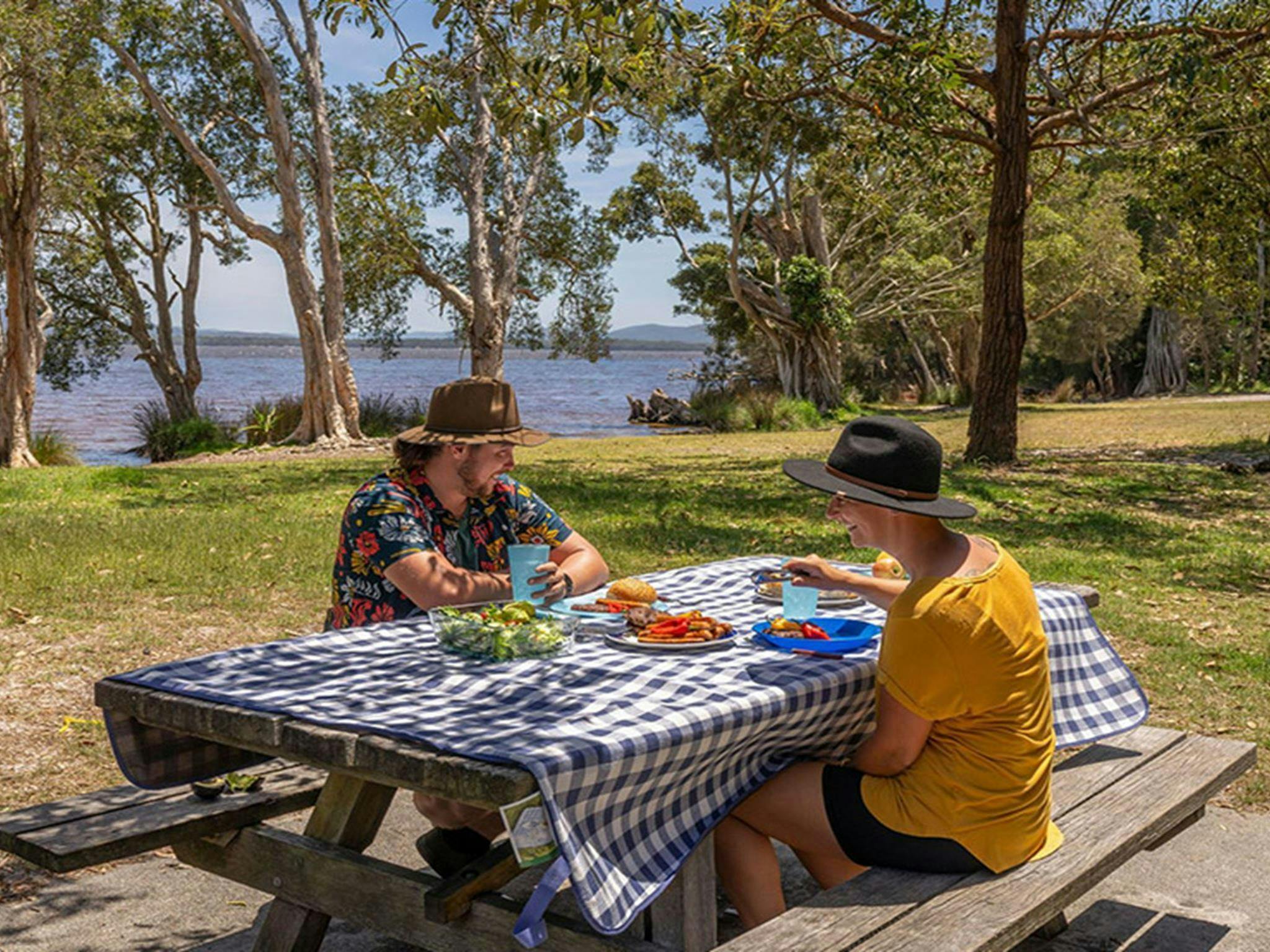 2 people having a picnic on a table by the water at Mungo Brush campground, Myall Lakes National