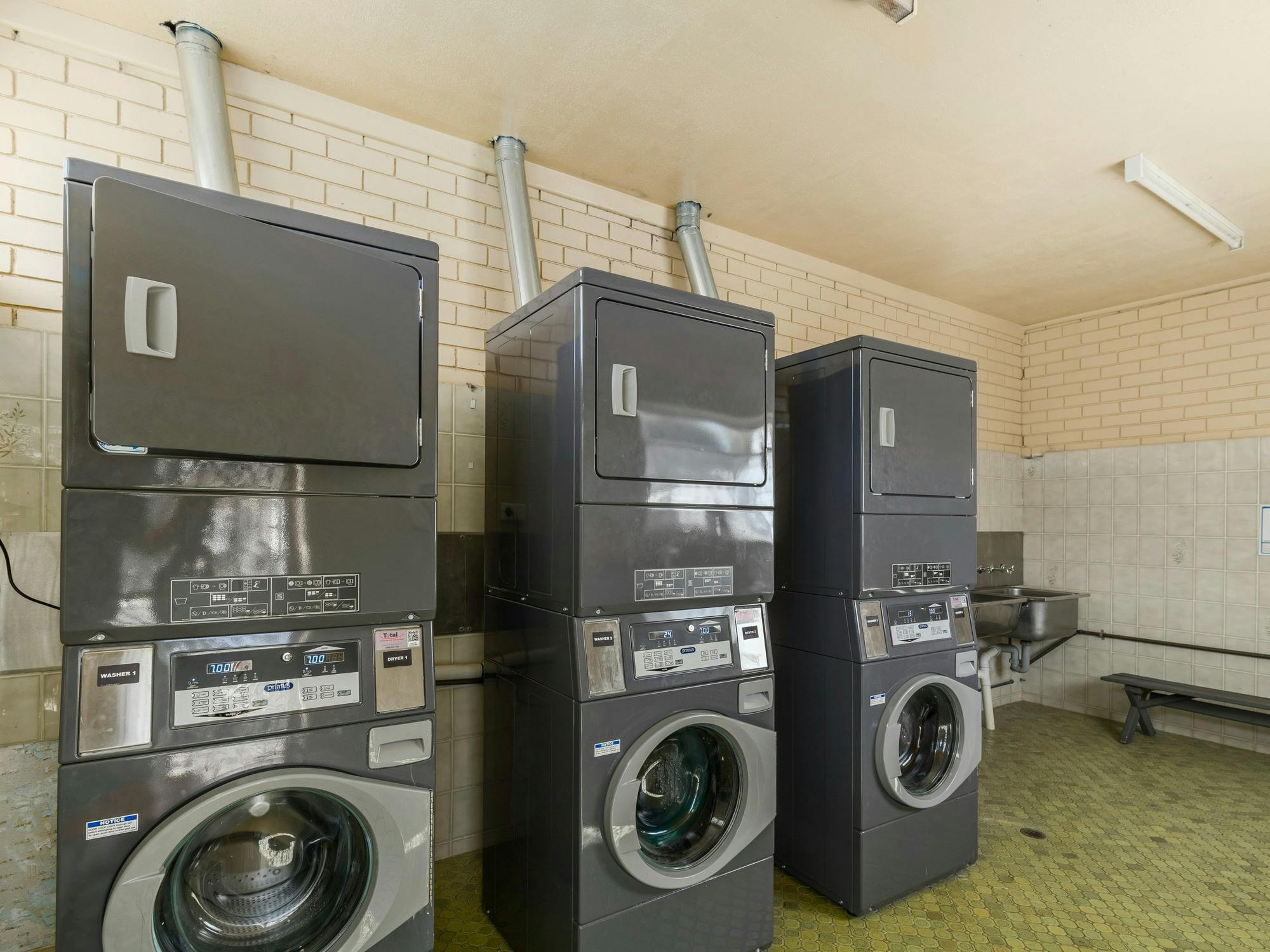 Guest laundry with large, modern washers and dryers up agains the wall, wooden bench and laundry tub