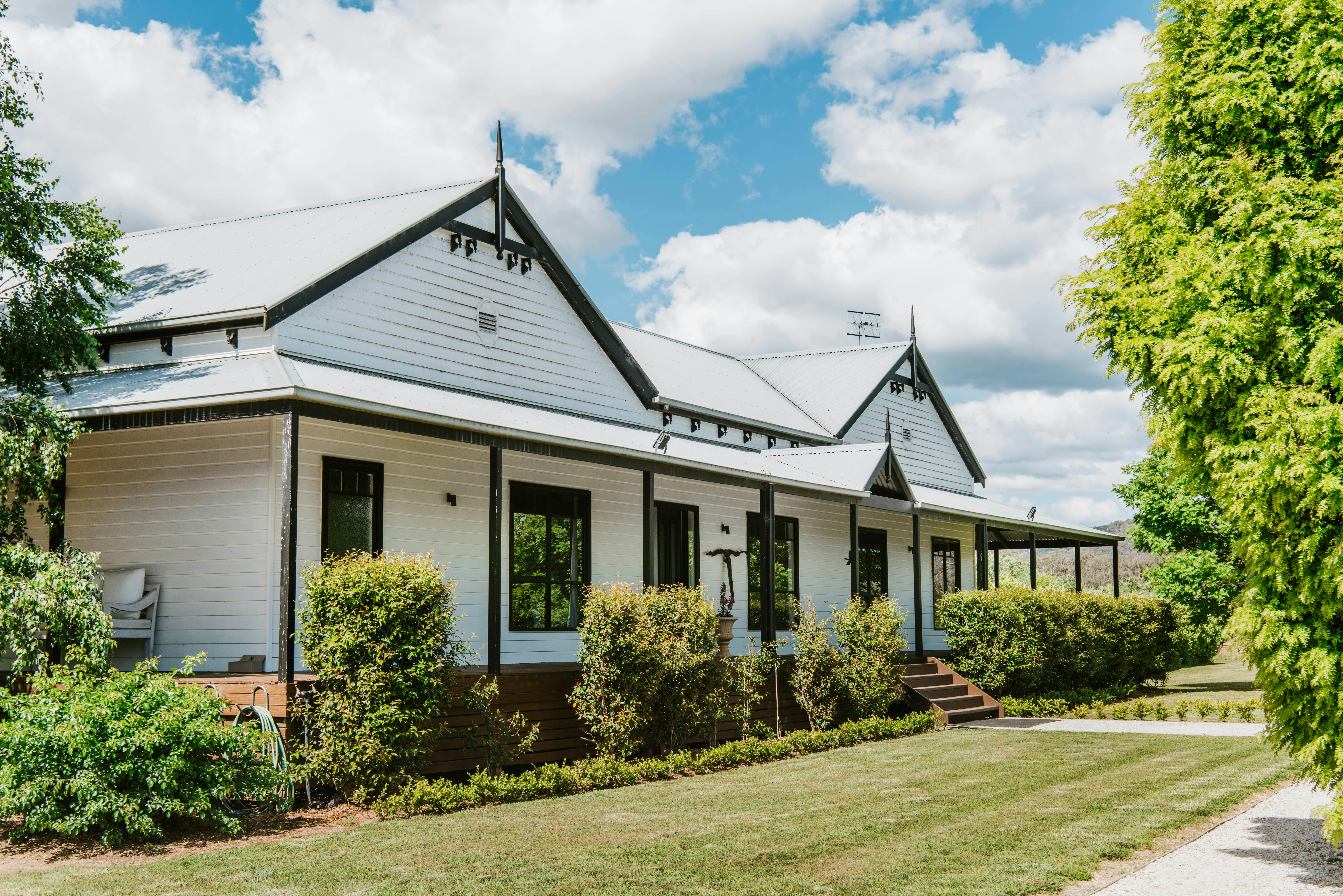 White farmhouse with gabled roof, black trim, and lush garden under a blue sky with clouds.