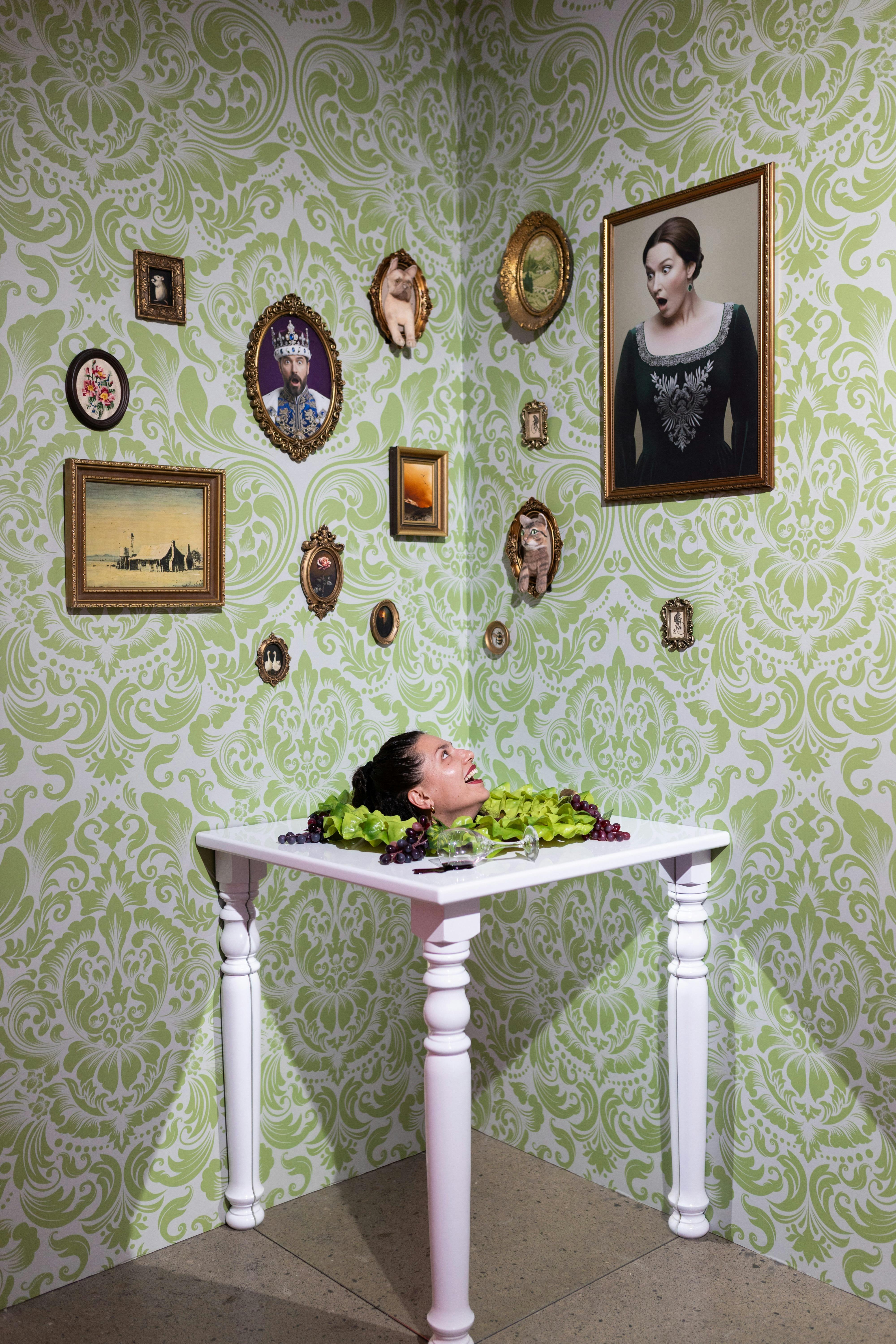 A surreal scene with a woman's head on a white table surrounded by grapes and lettuce