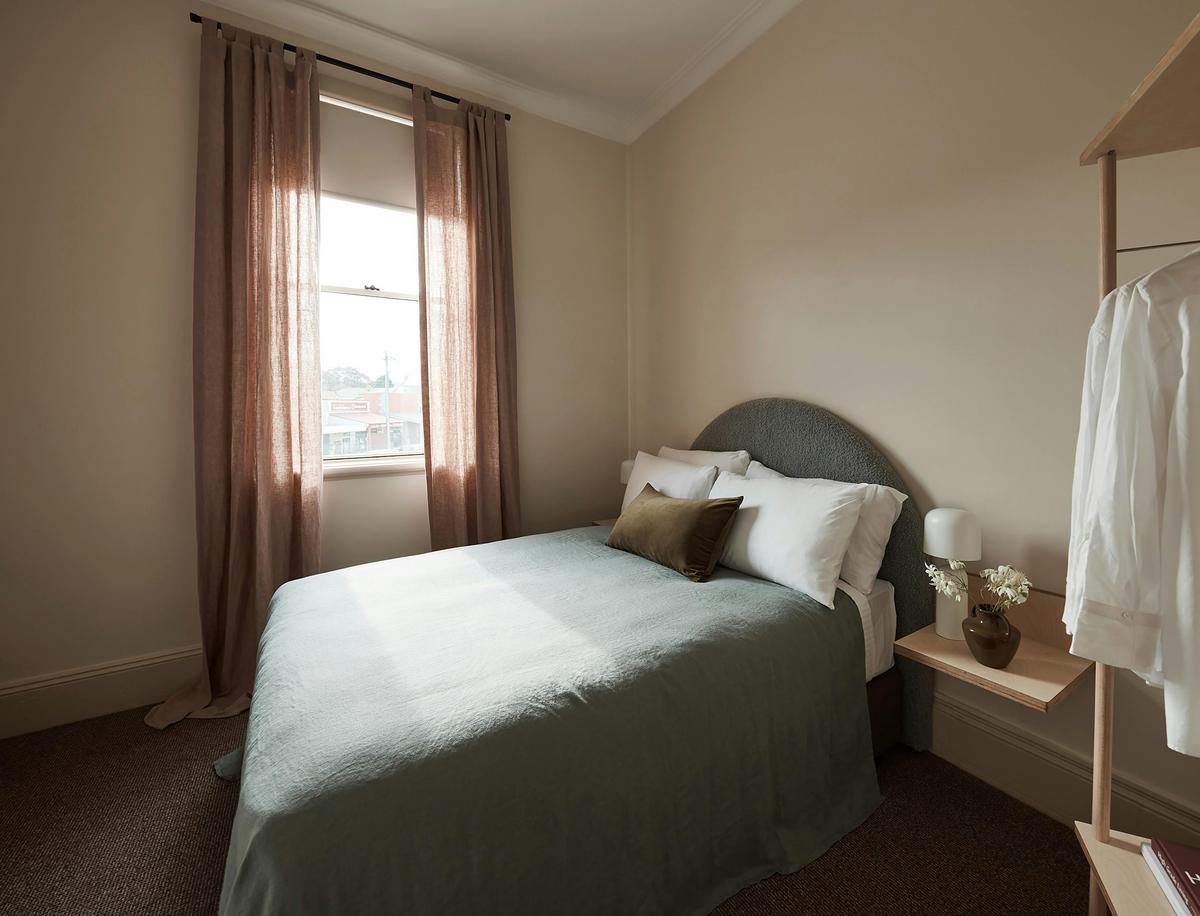 Bright bedroom with soft pink curtains, grey upholstered headboard, and green bedspread