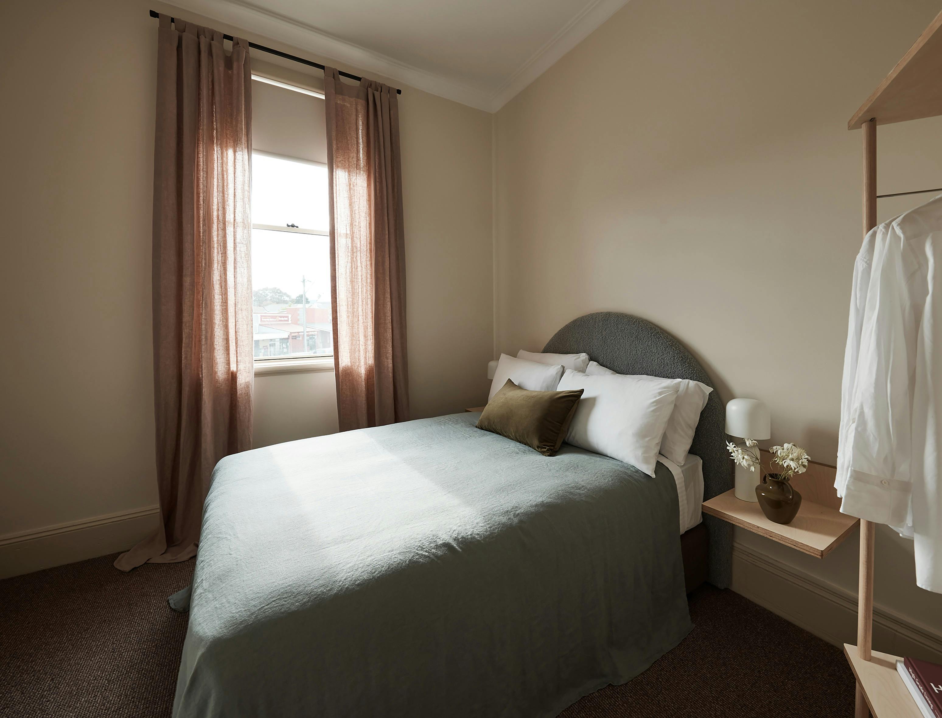 Bright bedroom with soft pink curtains, grey upholstered headboard, and green bedspread