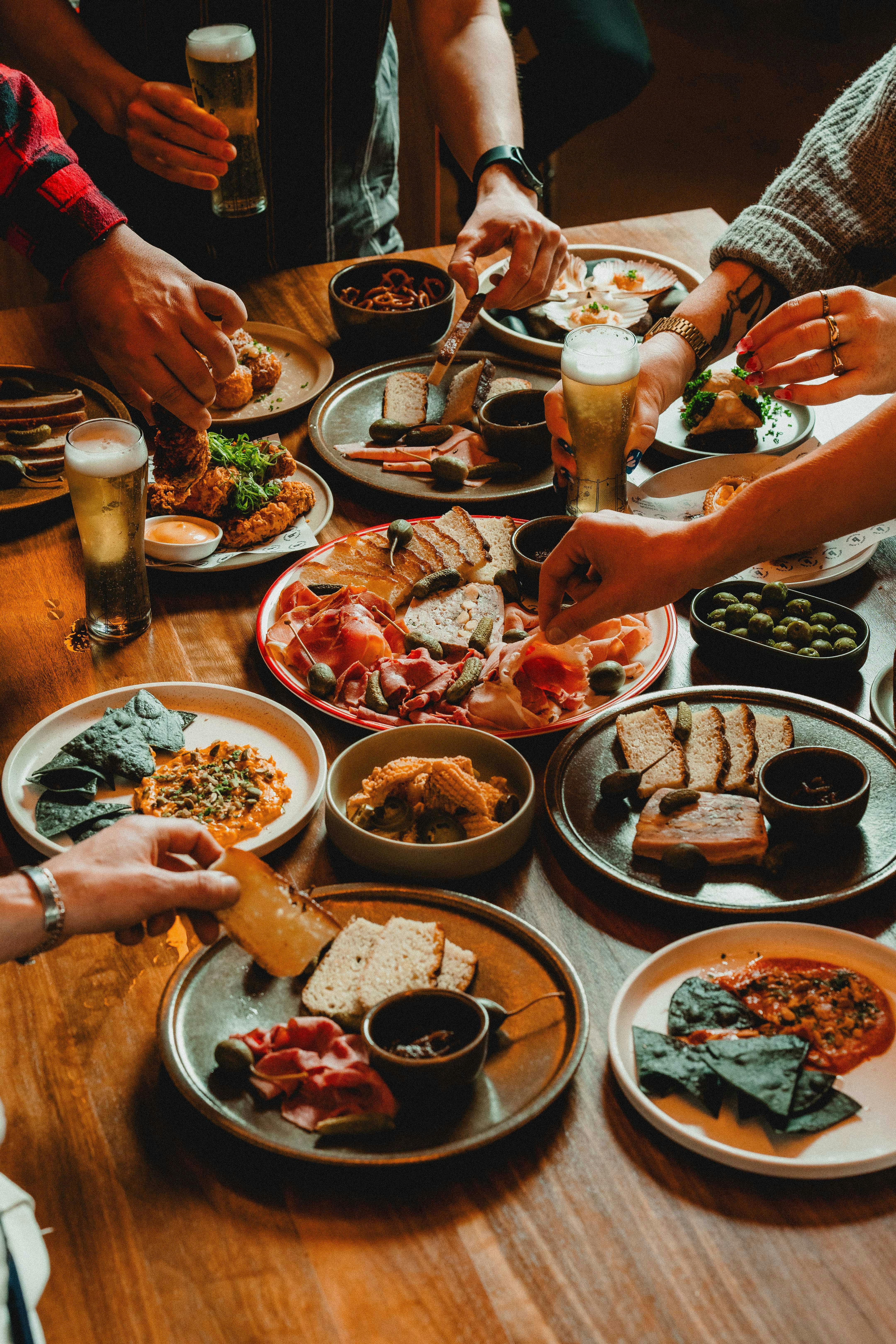 A table filled with shared plates of bread, meats, and snacks, with people reaching for food