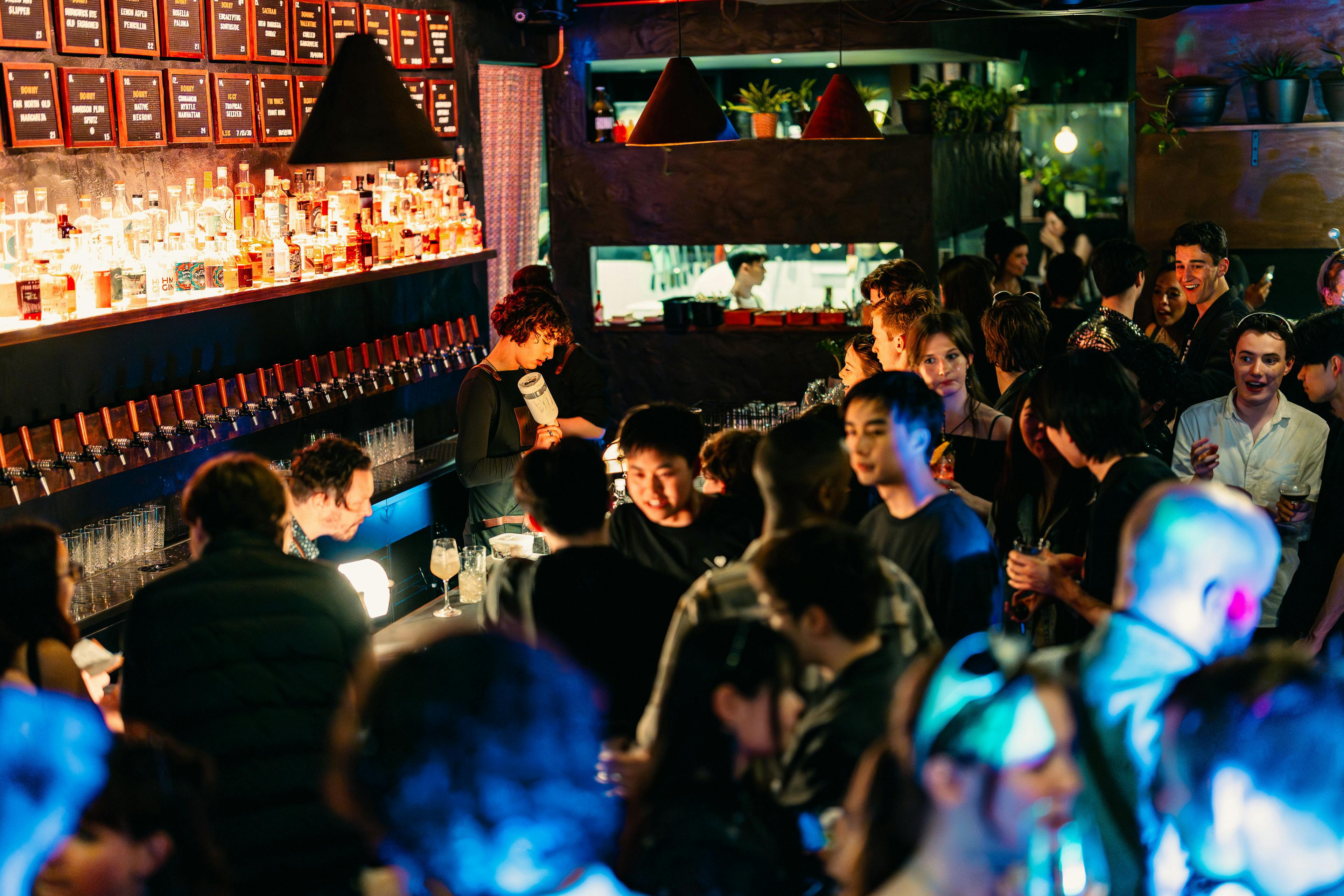 A group of young adults in a busy bar. A bartender in the background pours a drink.