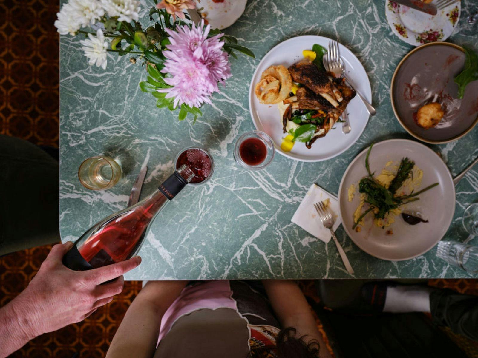 Plates of food on a laminex table and a hand pouring a glass of wine