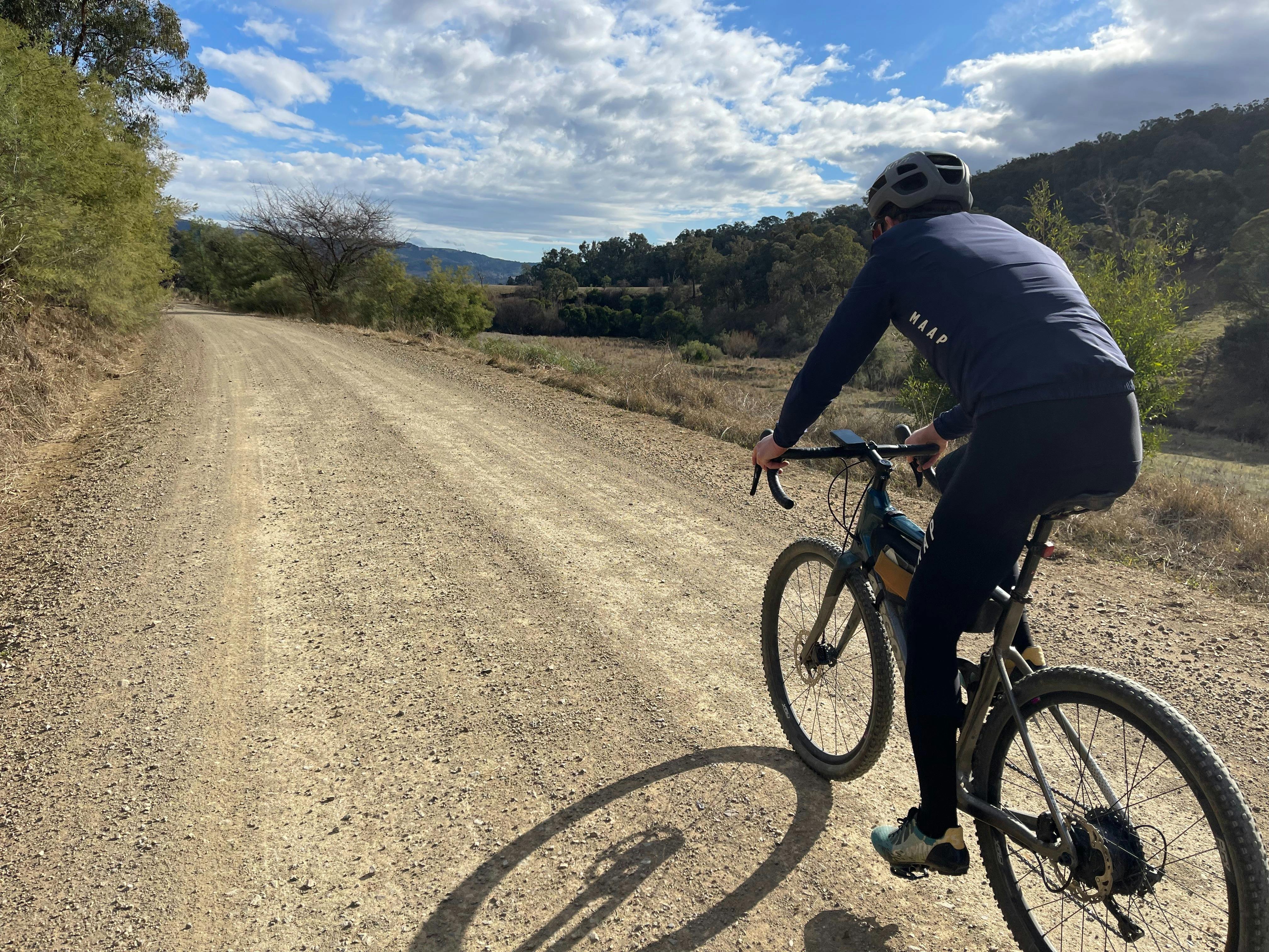 Lone cyclist riding away from camera on flat section of dirt road, hills and trees surrounding.