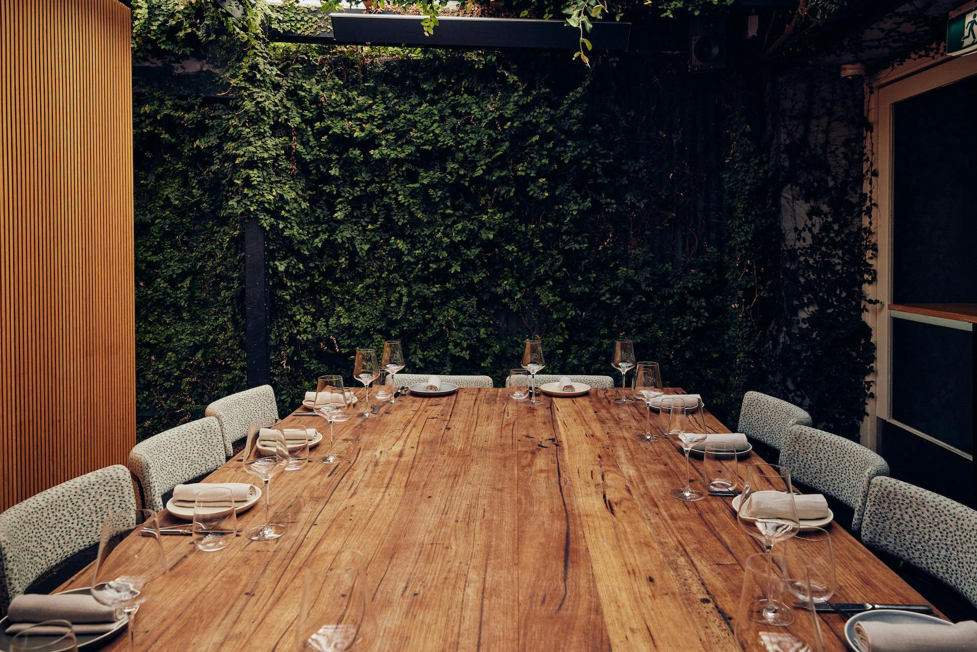 A large wooden table set for a group or function alongside a wall of greenery