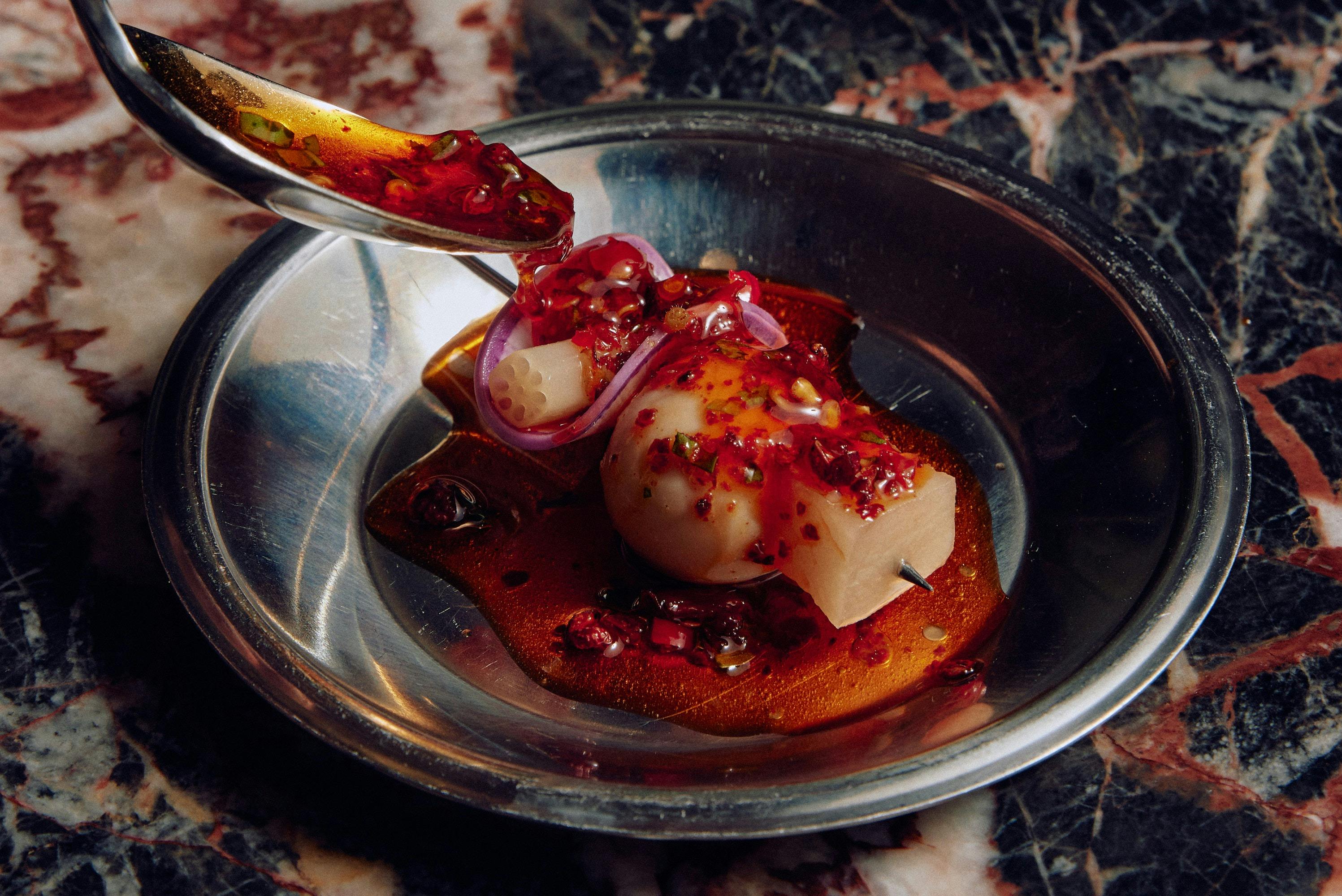 A spoon drips chilli oil onto a skewered egg, on a silver plate on top of a marble benchtop