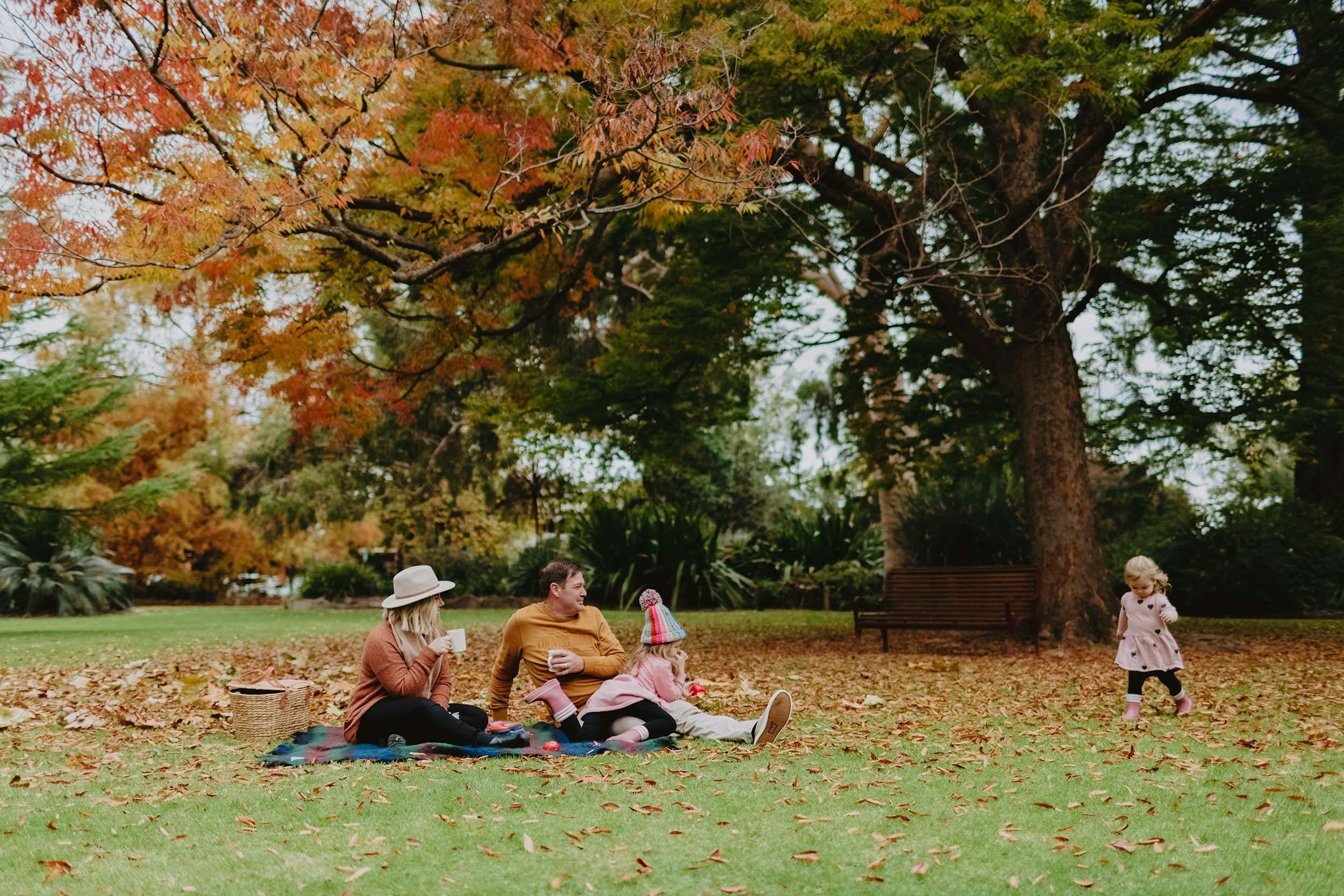 A family sit on a picnic rug underneath autumn trees at the Albury Botanic Gardens. A little girl in