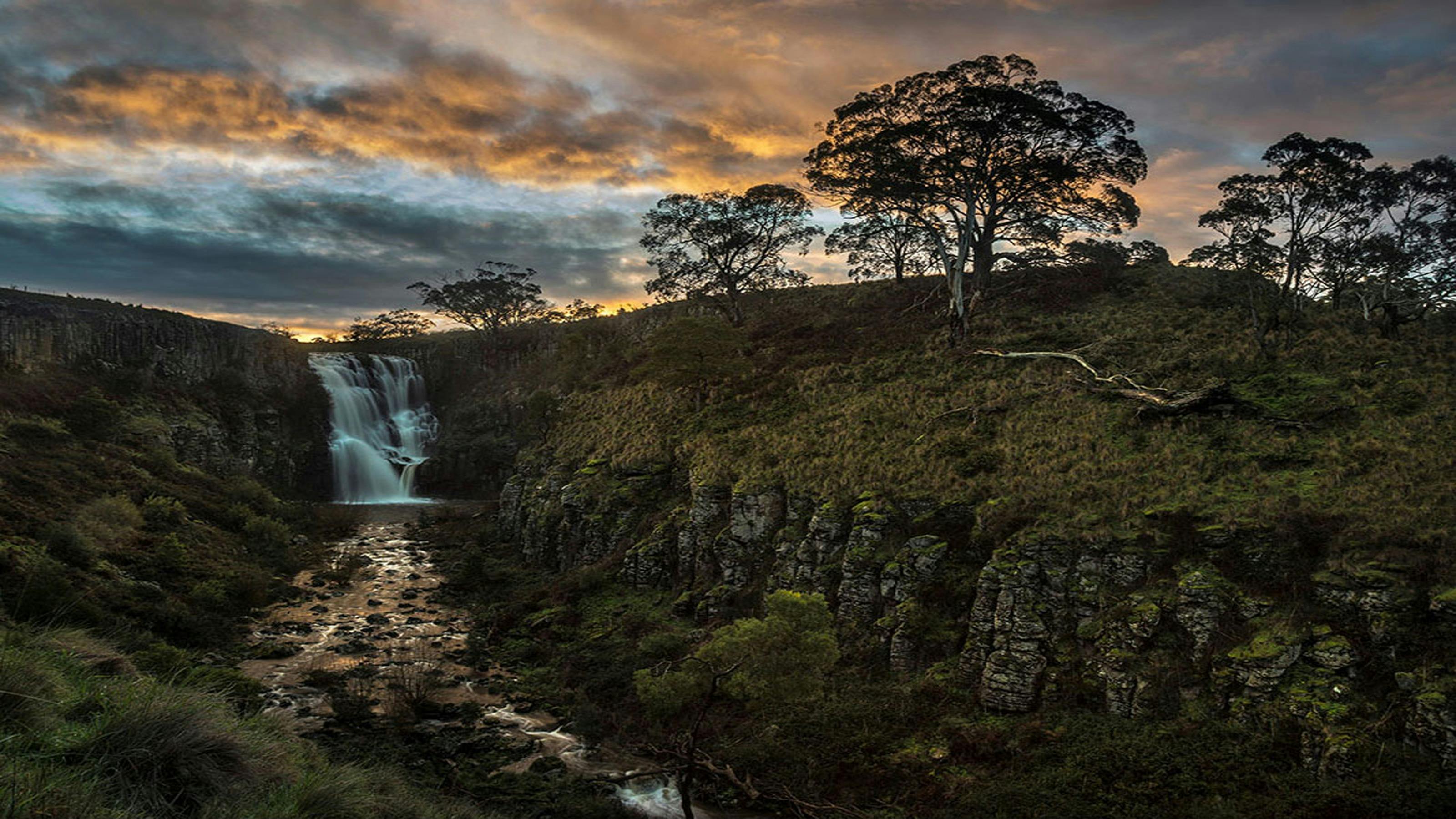wide angle of waterfall with moody grey and orange sky and cliff with moss and trees