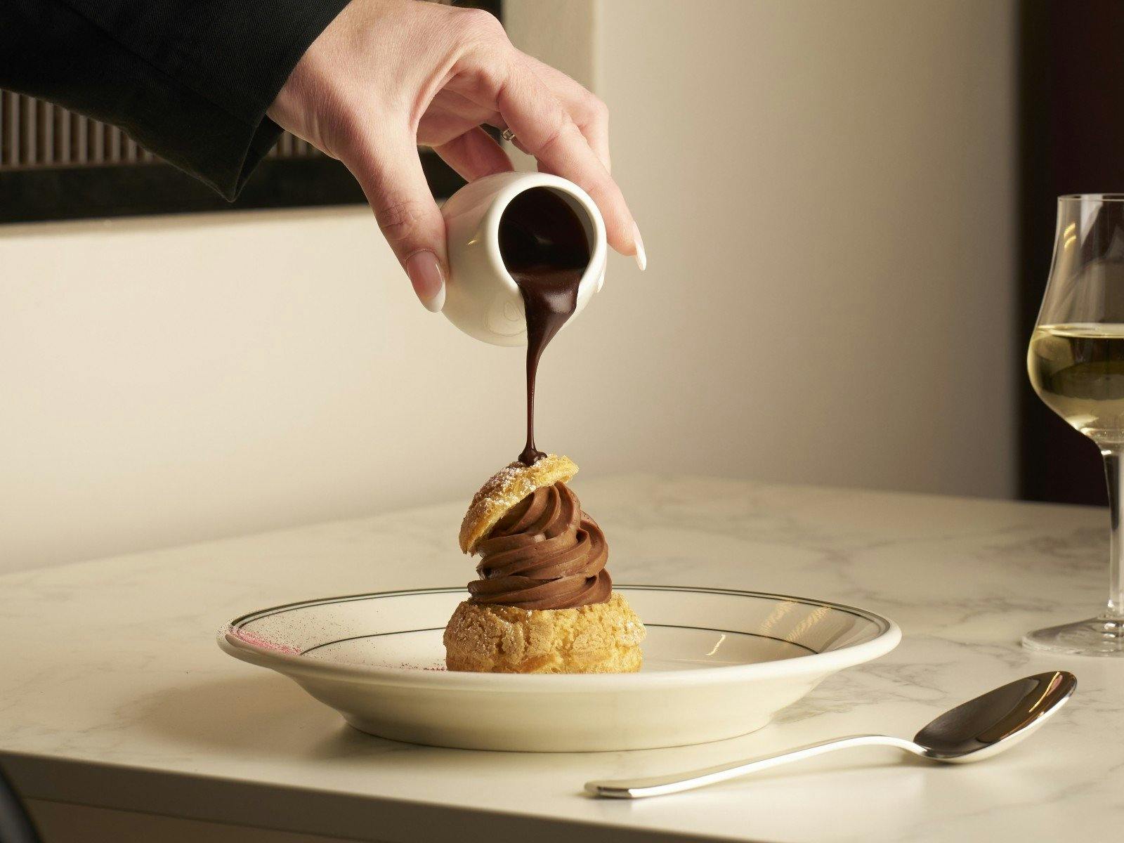 a pastry with chocolate cream in the middle while a hand pours chocolate sauce on it