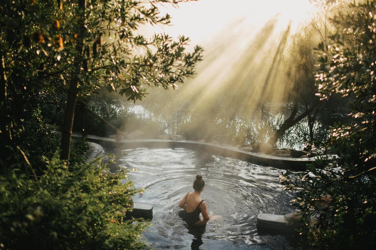 a woman in a hot spring surrounded by trees with sunlight streaming in