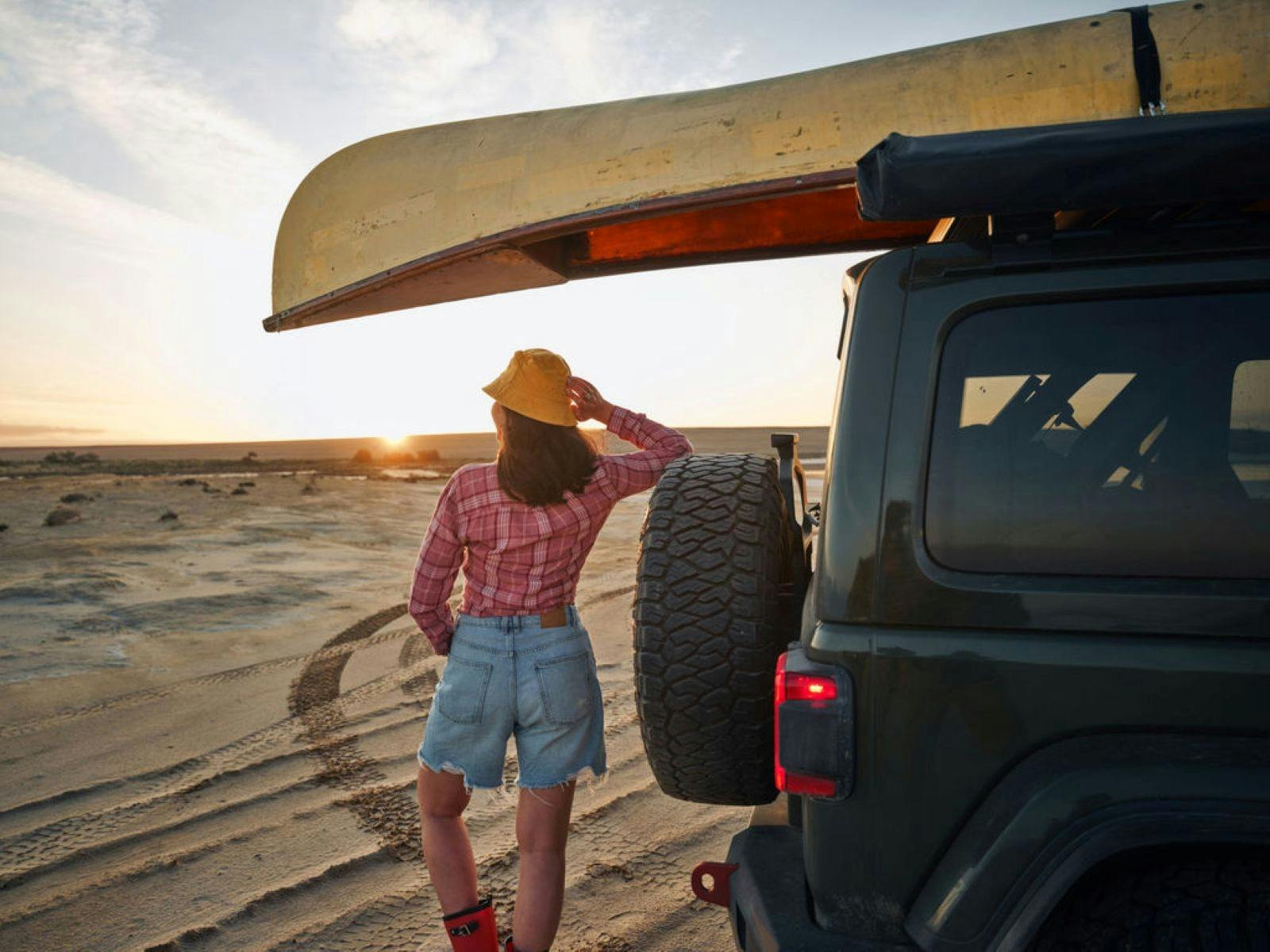 Person standing at the back of a small off road vehicle with a canoe on the roof looking at the sun