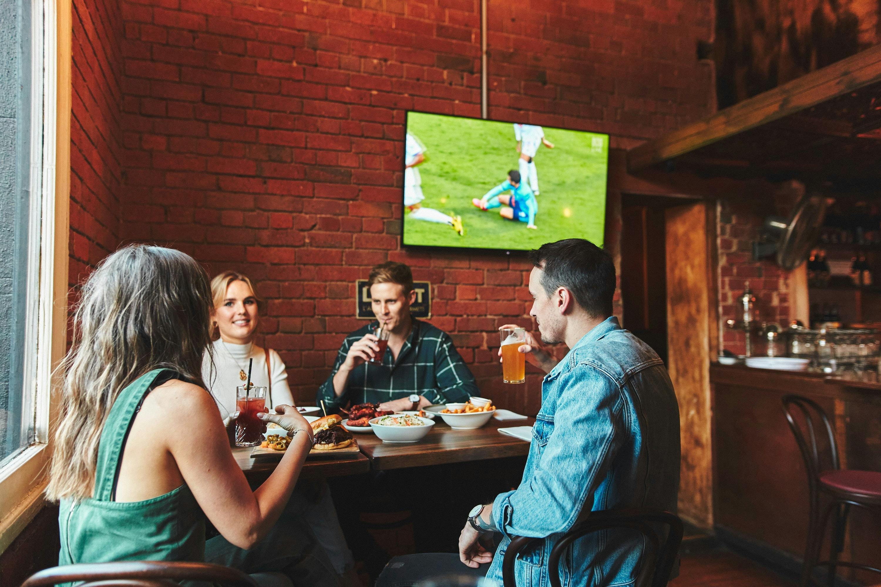 four people seated at a table and enjoying fod and drinks while watching a game on the television