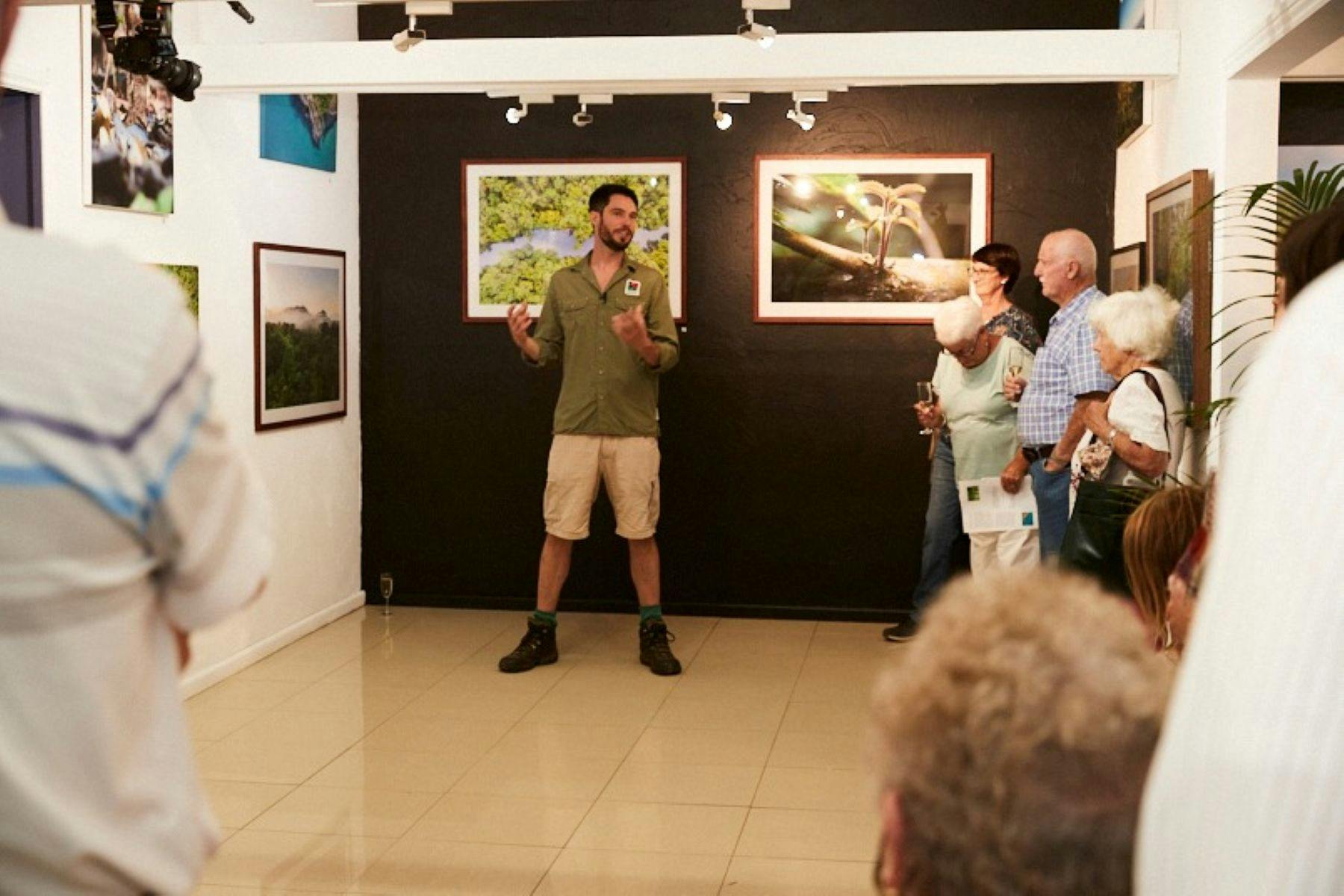 Person standing in front of artworks on a wall and talking to a group of people