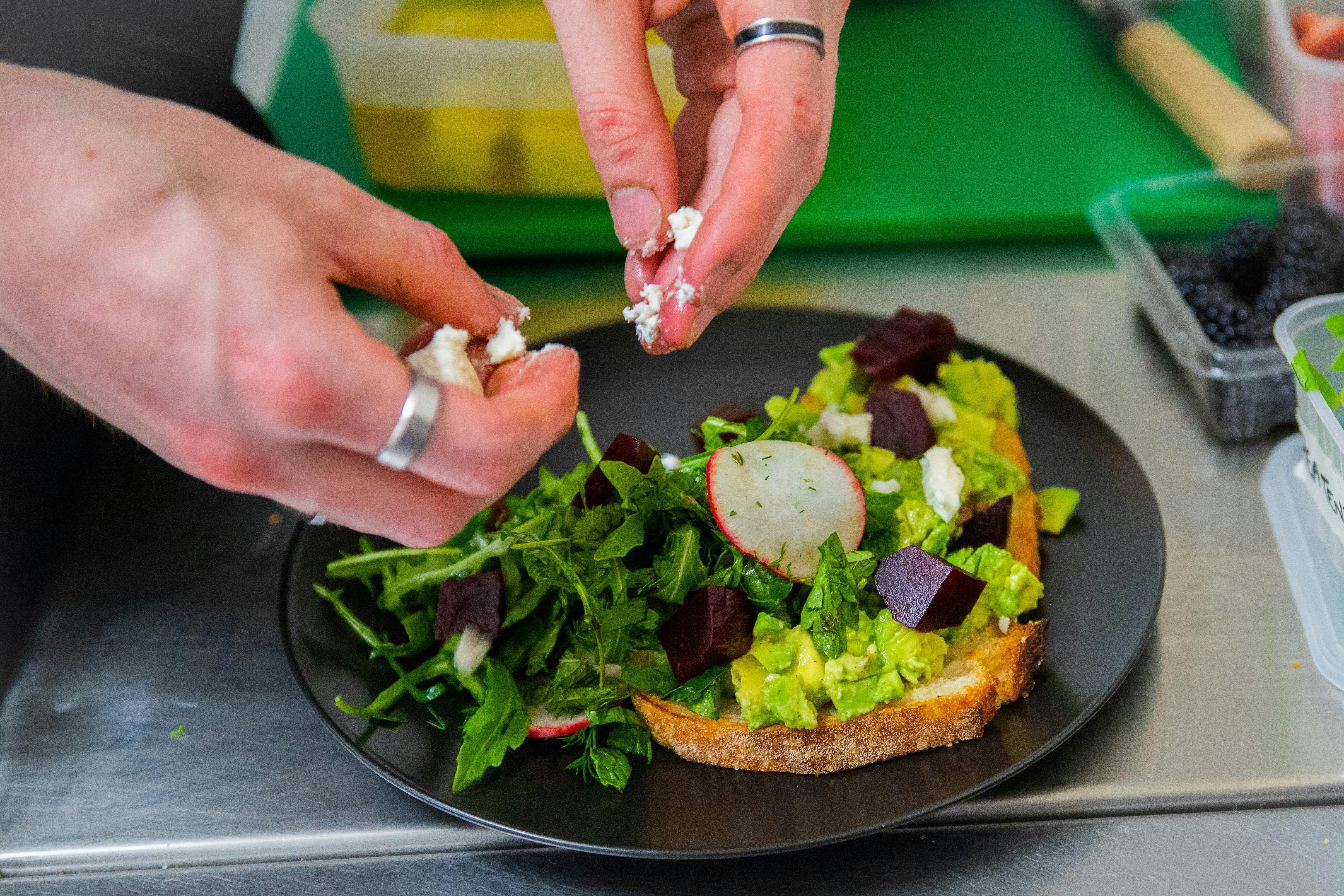 Smashed Avocado being plated in the kitchen with hands crumbling goat's cheese over the top