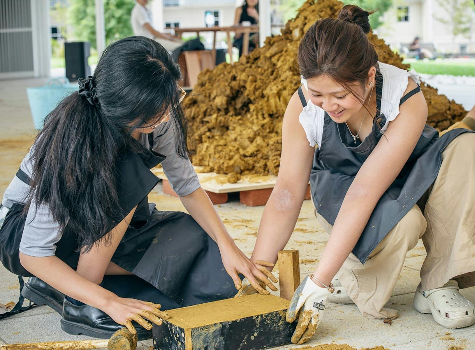 Two people building brick like structure with natural materials.