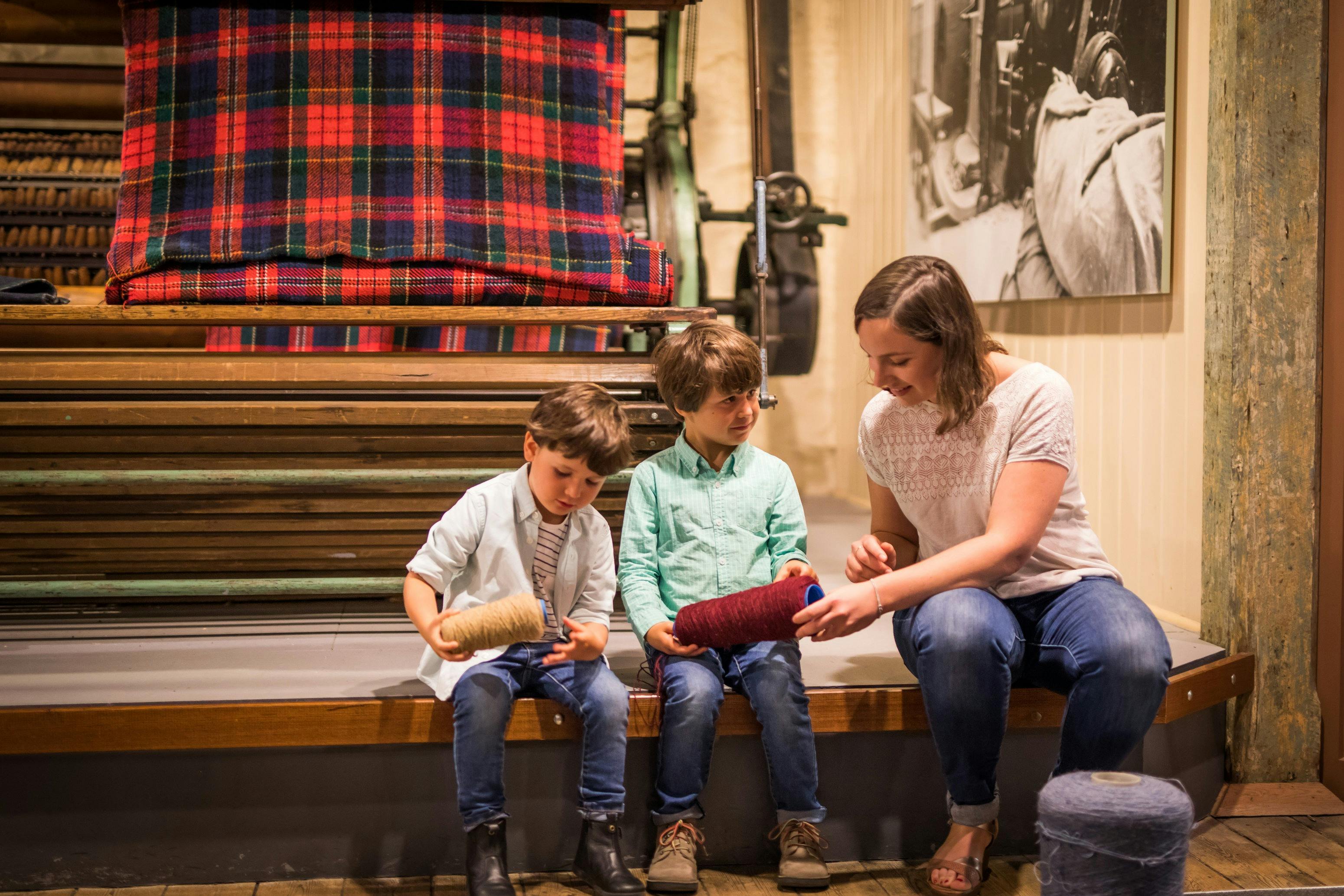 Two children holding two rolls of yarn and a woman seated with them at the National Wool Museum