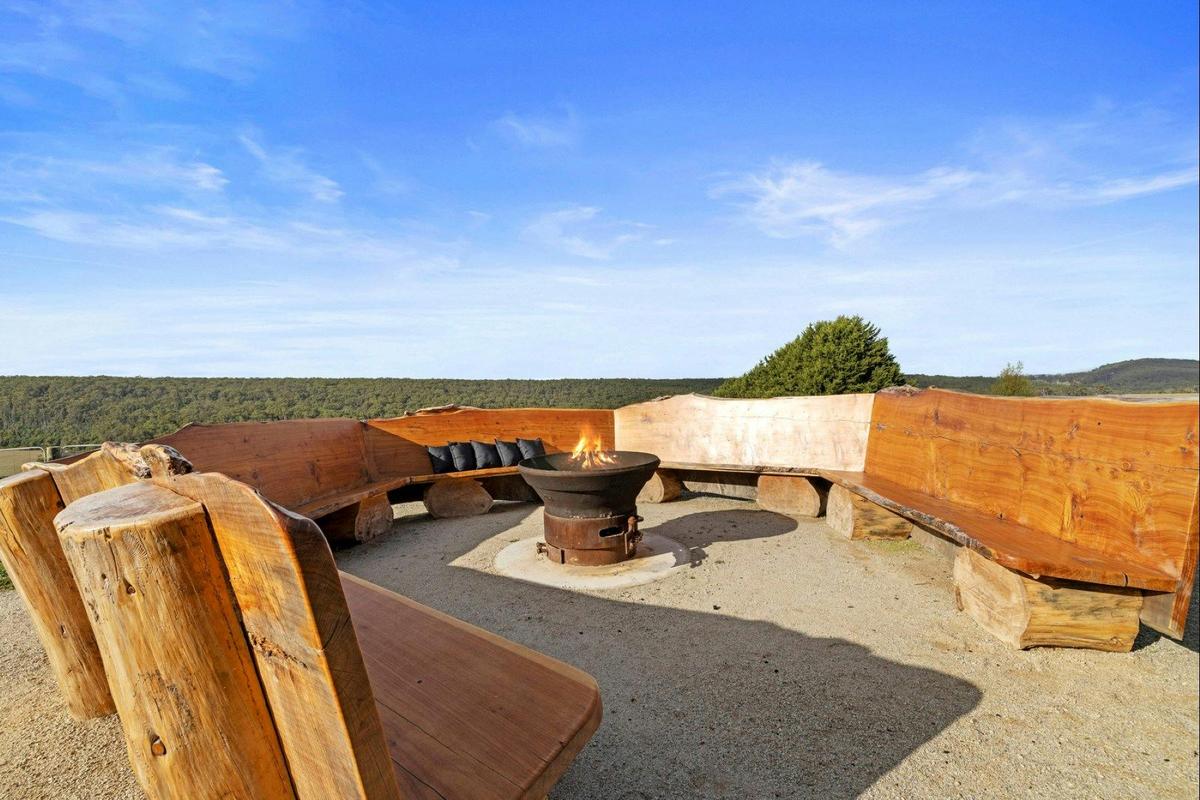an outdoor wooden bench curved around a fire pit with a lit fire with bushlands in the background