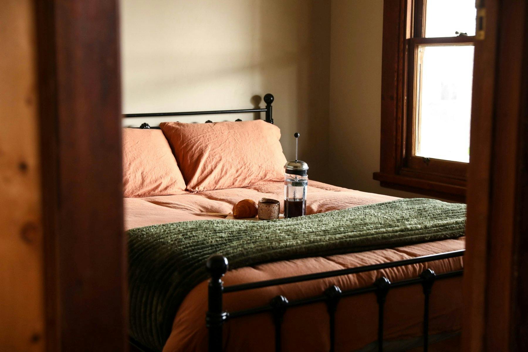 a double bed with pink linen and green throw with a coffee press and stone cup