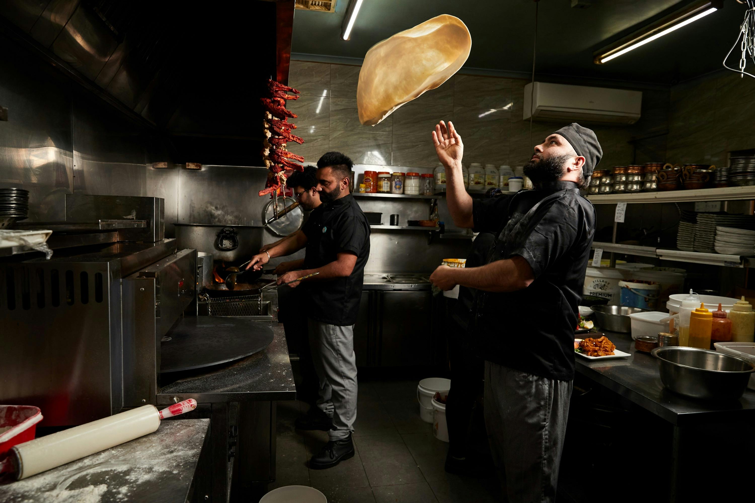 Three chefs cooking in a kitchen with one of them throwing naan bread in the air.