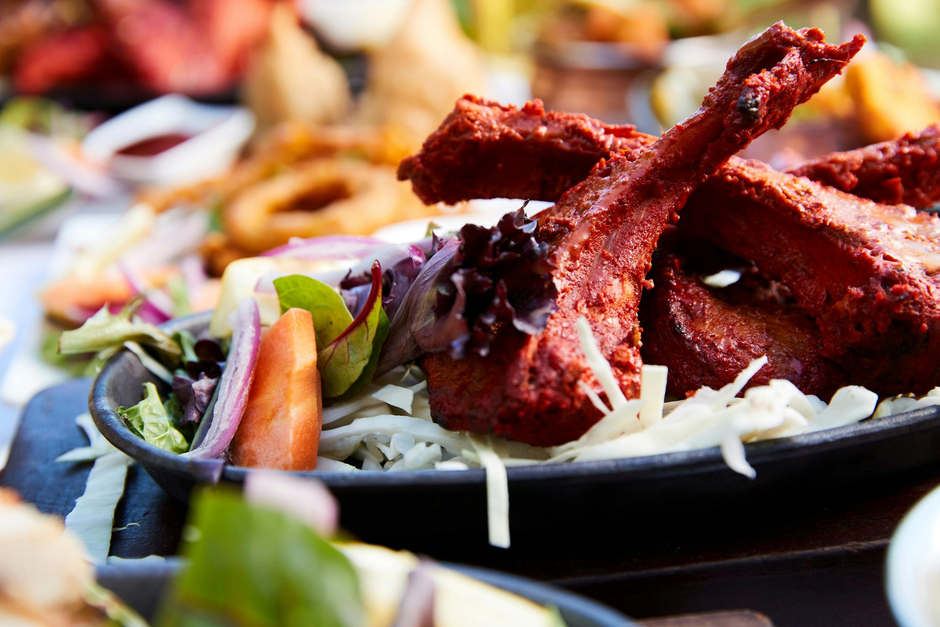A platter of dark red tandoori lamb with fresh vegetables around it.