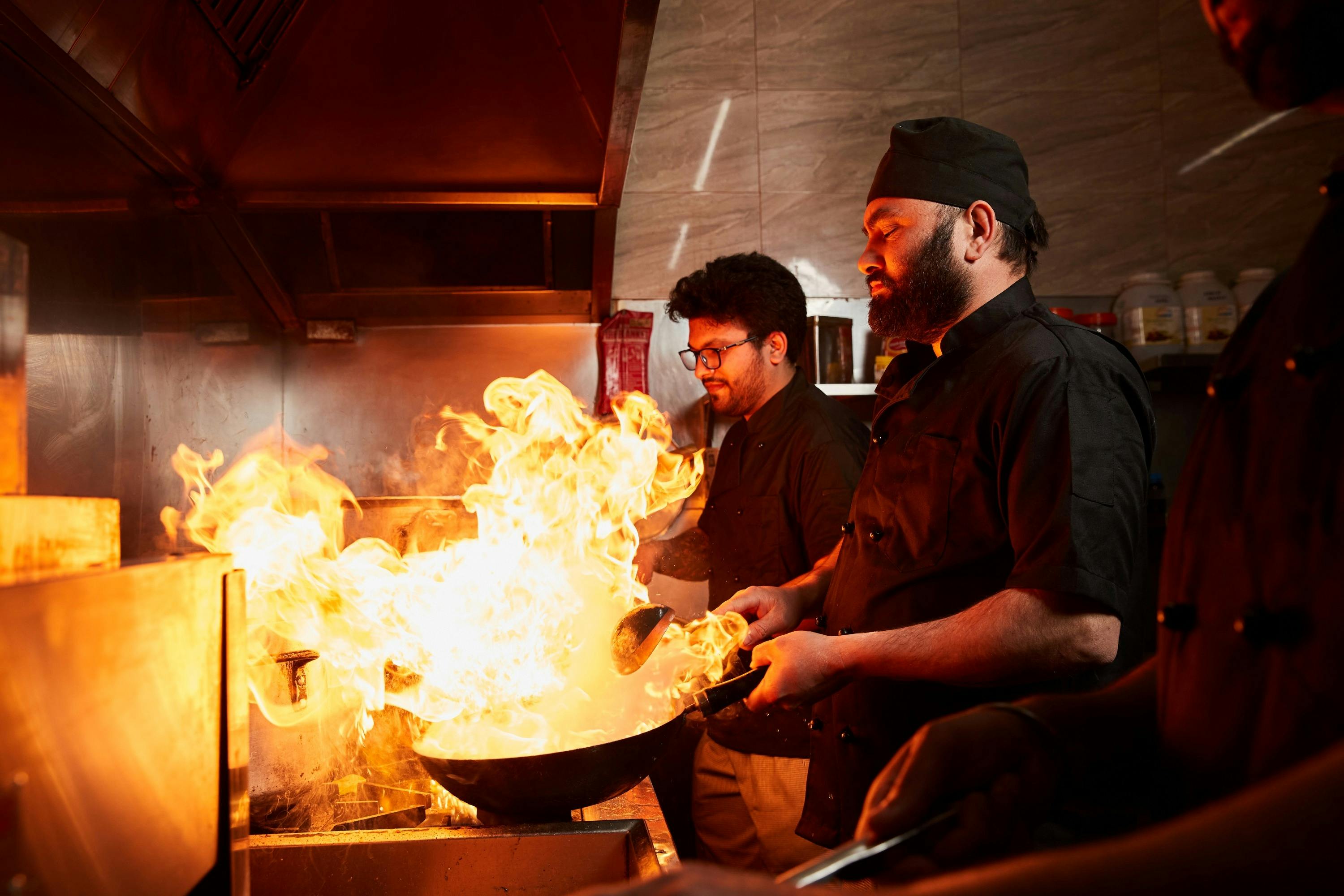 Three chefs cooking next to each other in a kitchen with flames erupting from a frying pan.