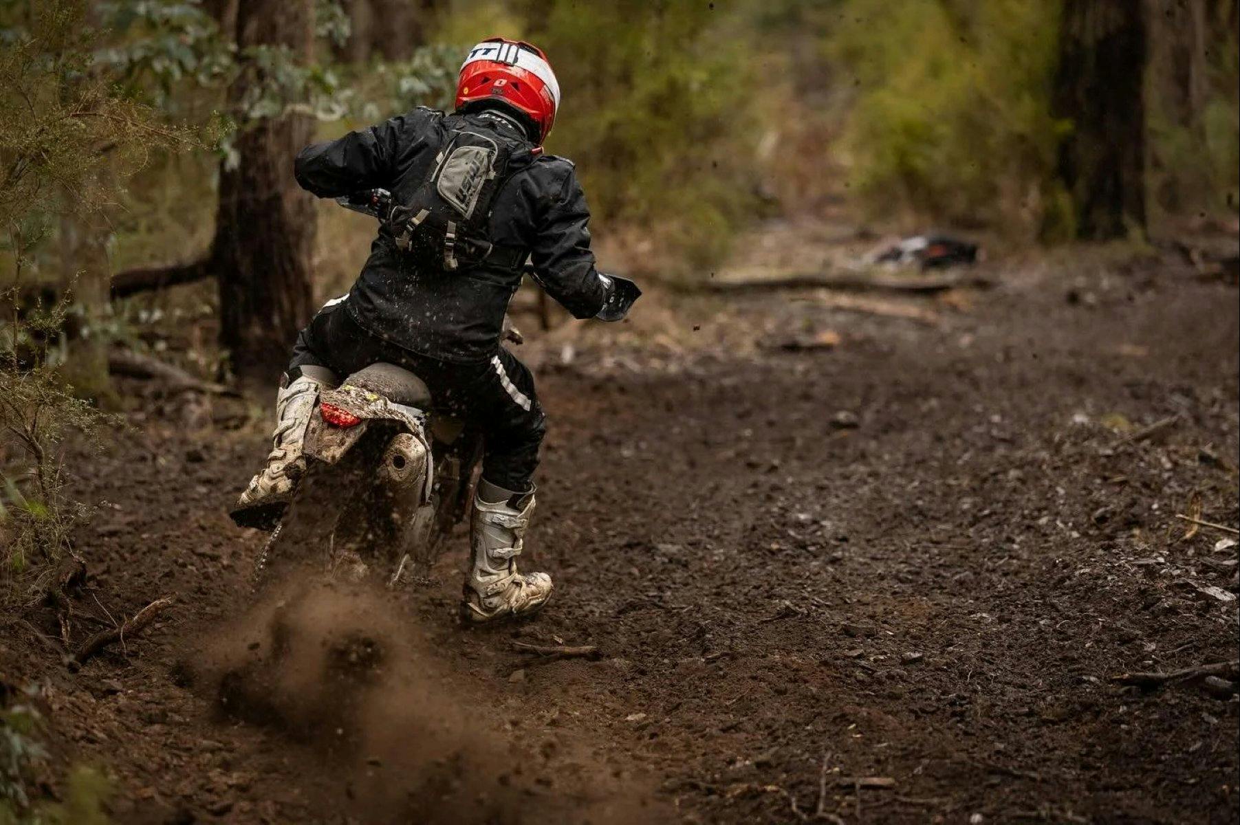 person in a black race suit with a helmet riding a dirt bike in the forest