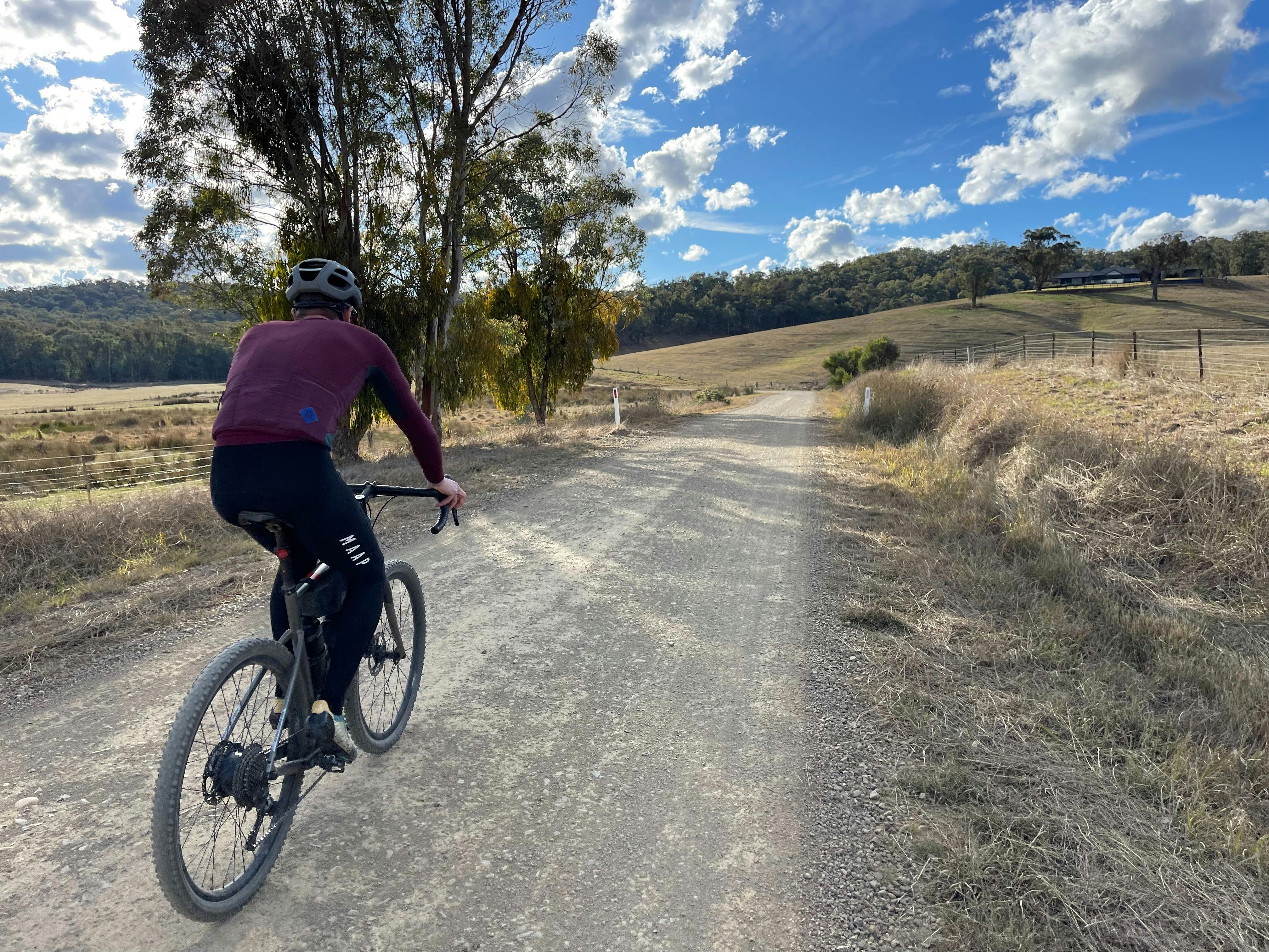 Clist riding away from camera towards bend in gravel road, hills, farmland and house in distance.