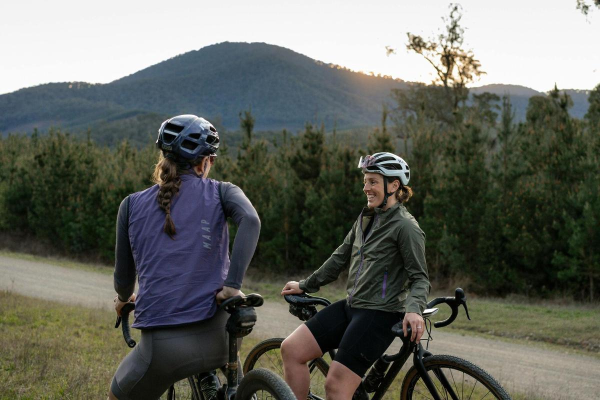 Two cyclists taking a break from riding on a gravel road with bushland and hills surrounding them.