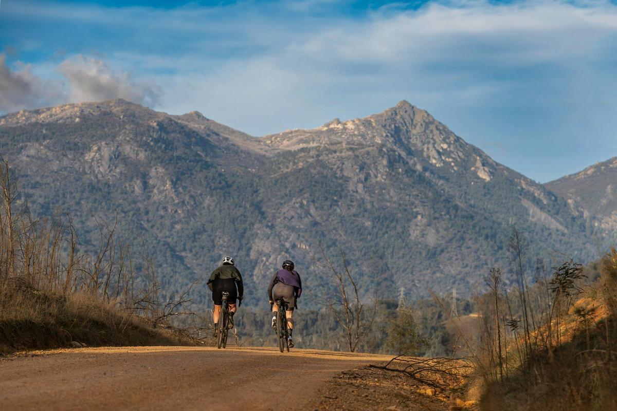 The back of two people on bikes riding on a gravel road with large mountain in backdrop.